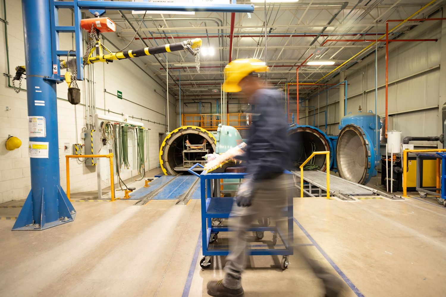 Worker in yellow hard hat pushing cart through industrial manufacturing facility with large blue machinery.