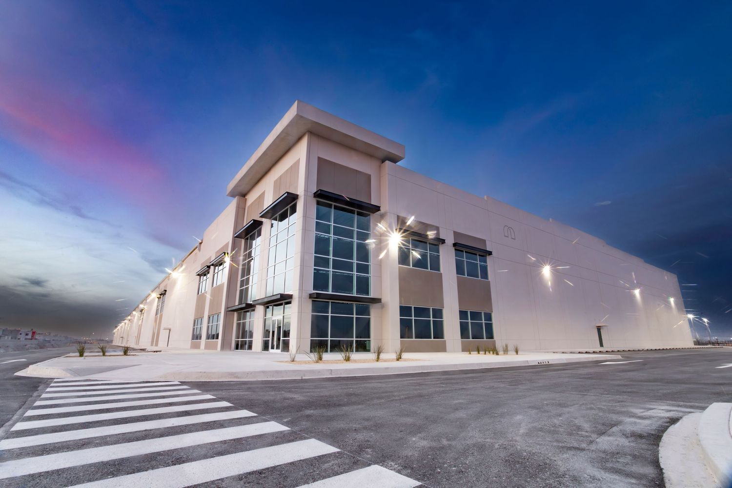 Modern large warehouse building exterior at dusk with illuminated facade, parking lot, and vibrant blue sky.
