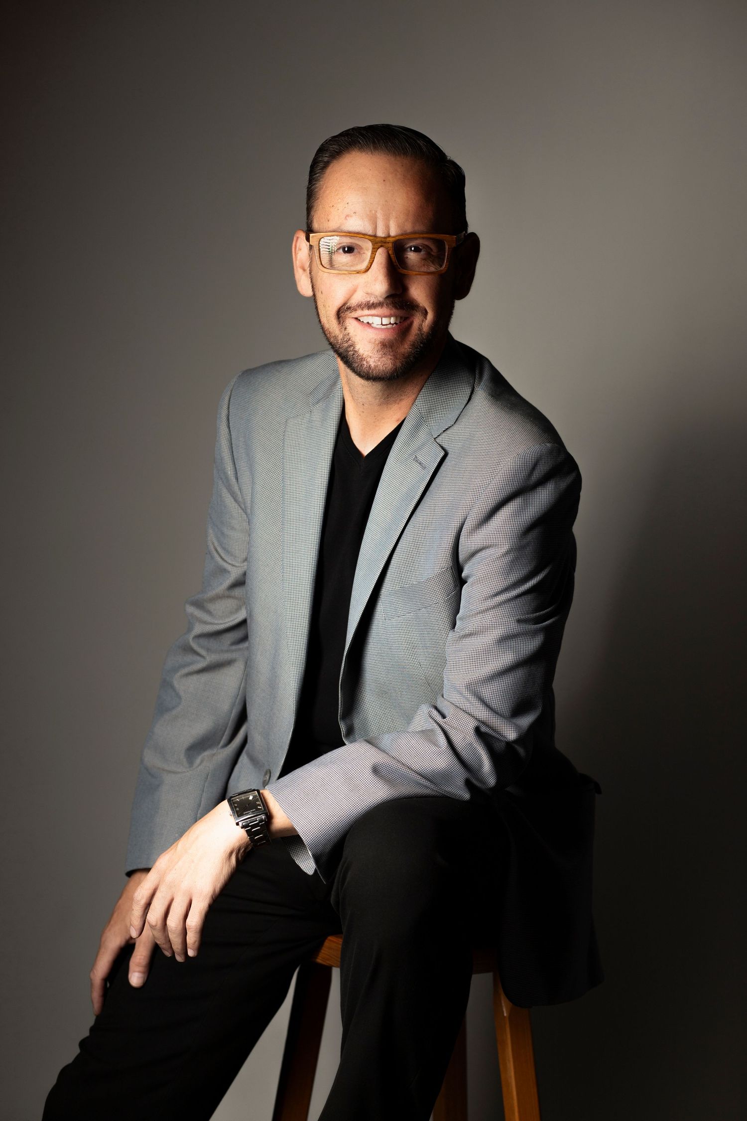 Professional studio portrait of someone wearing a gray blazer and black shirt against dark background.