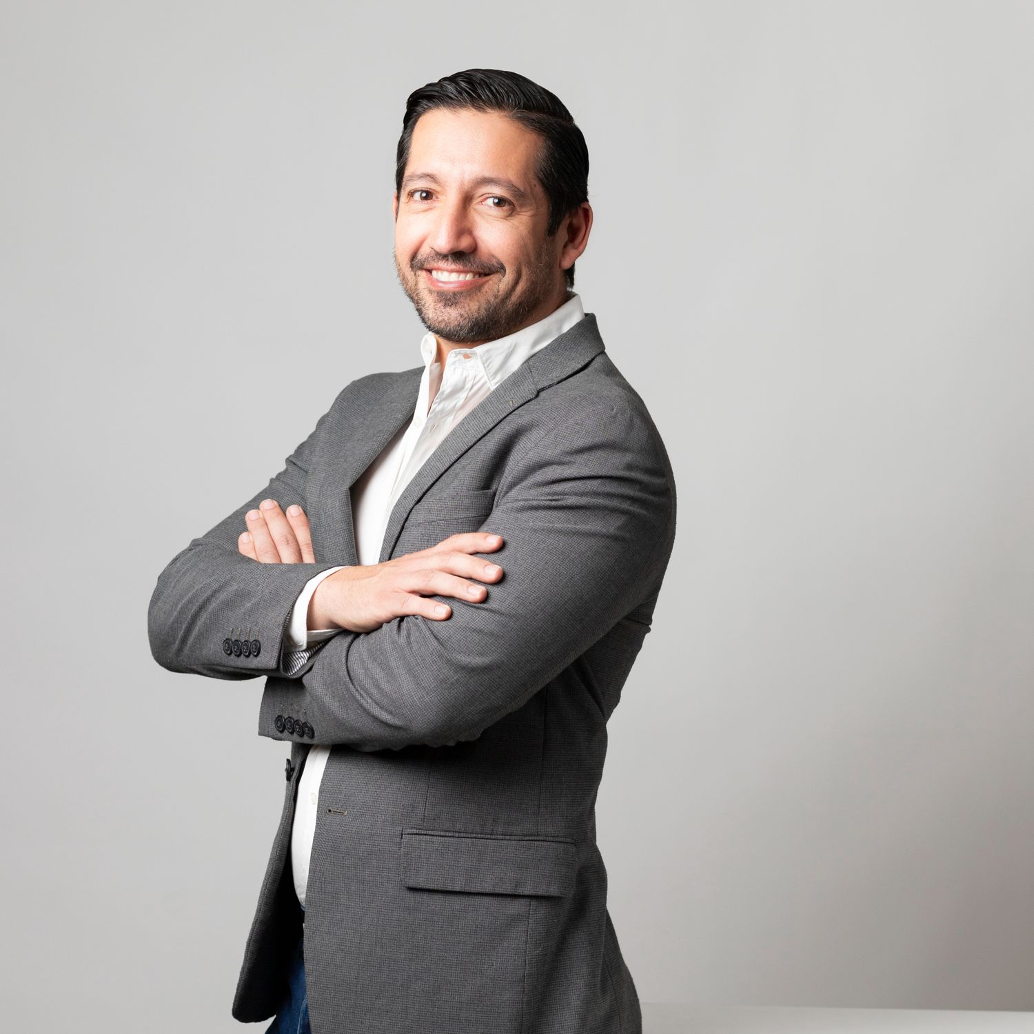 Professional portrait of a person in a grey suit and white shirt smiling confidently with arms crossed.