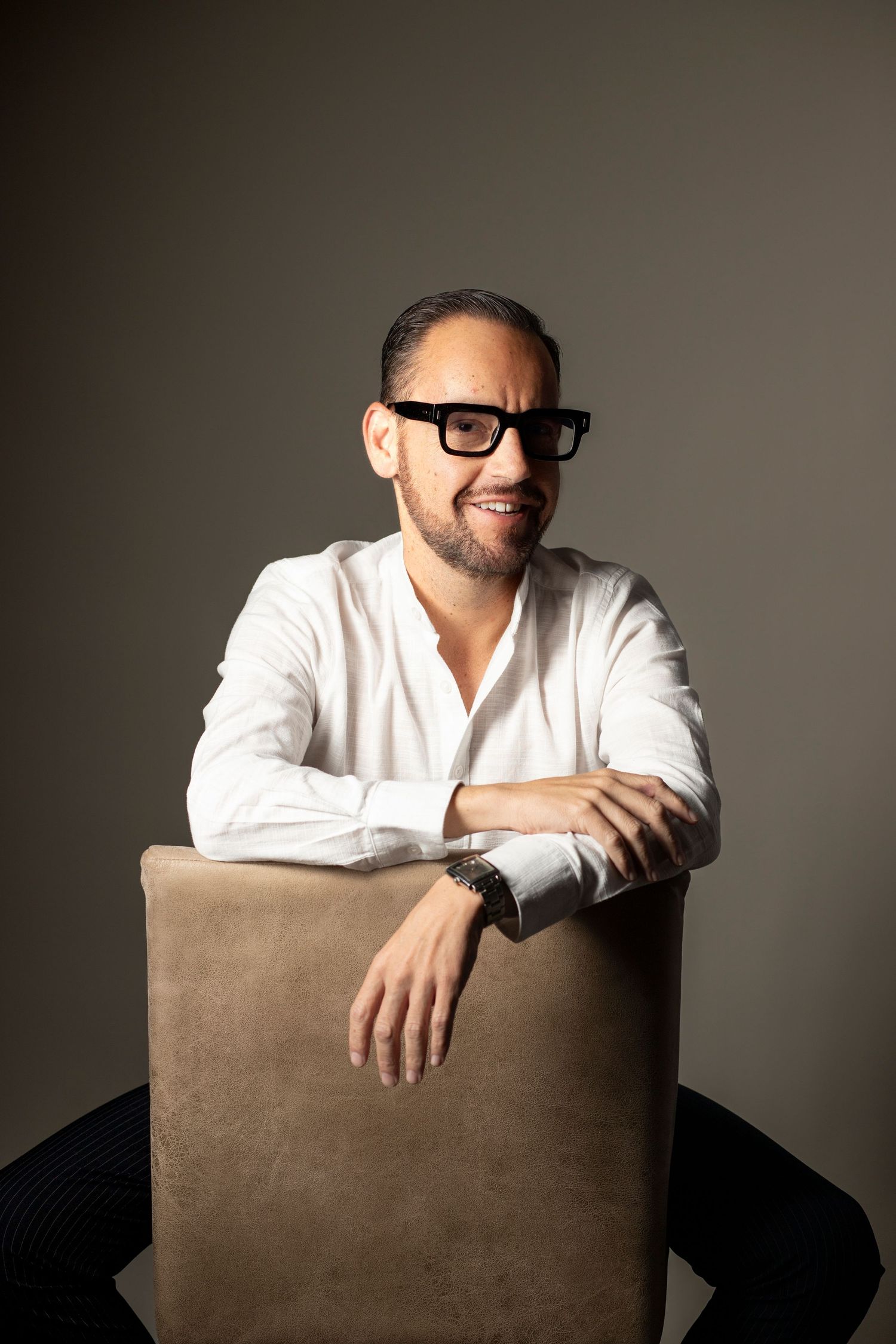Studio portrait showing someone in white shirt leaning on brown chair with dramatic lighting.
