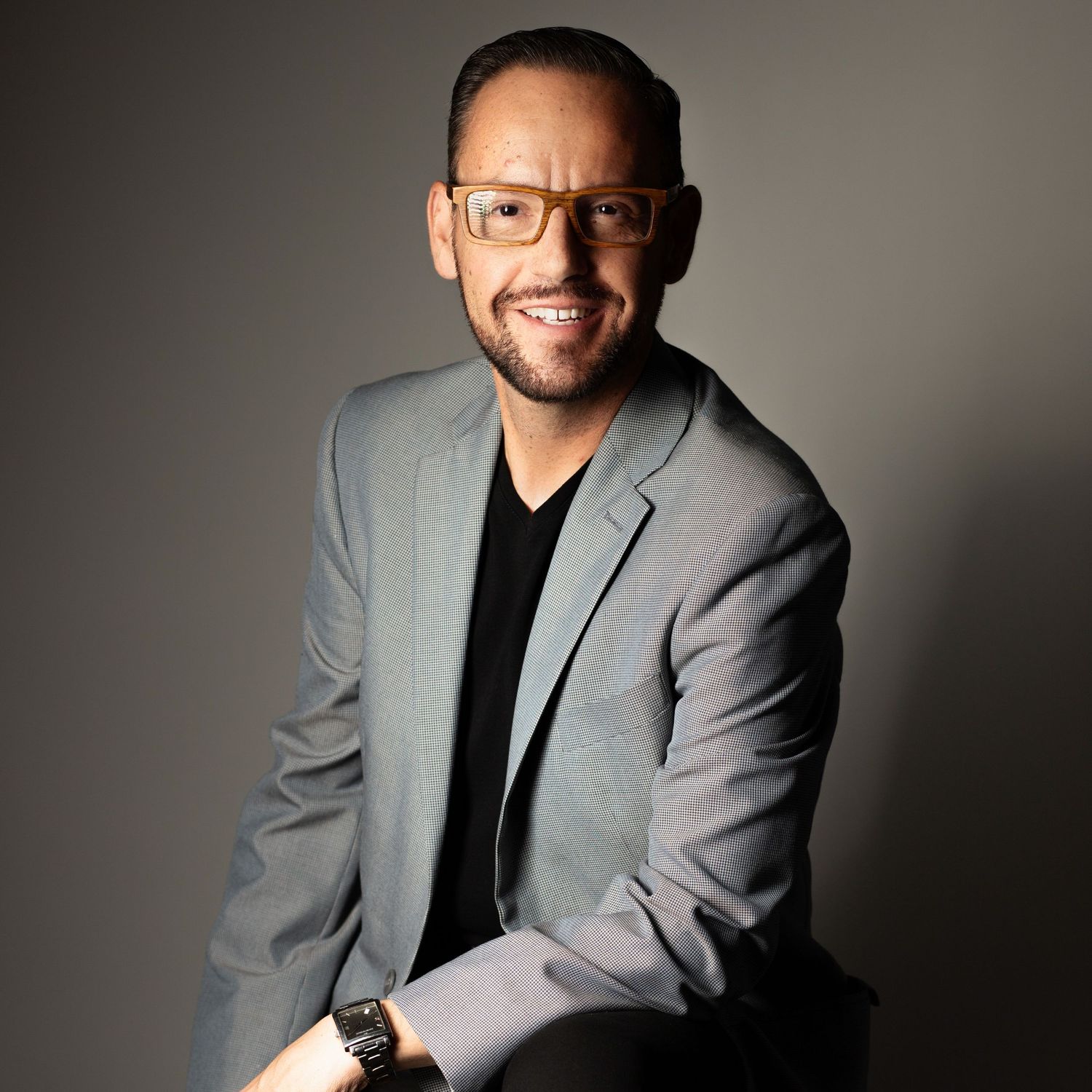 Professional headshot of someone wearing glasses and a gray blazer over black shirt against neutral background, smiling warmly.