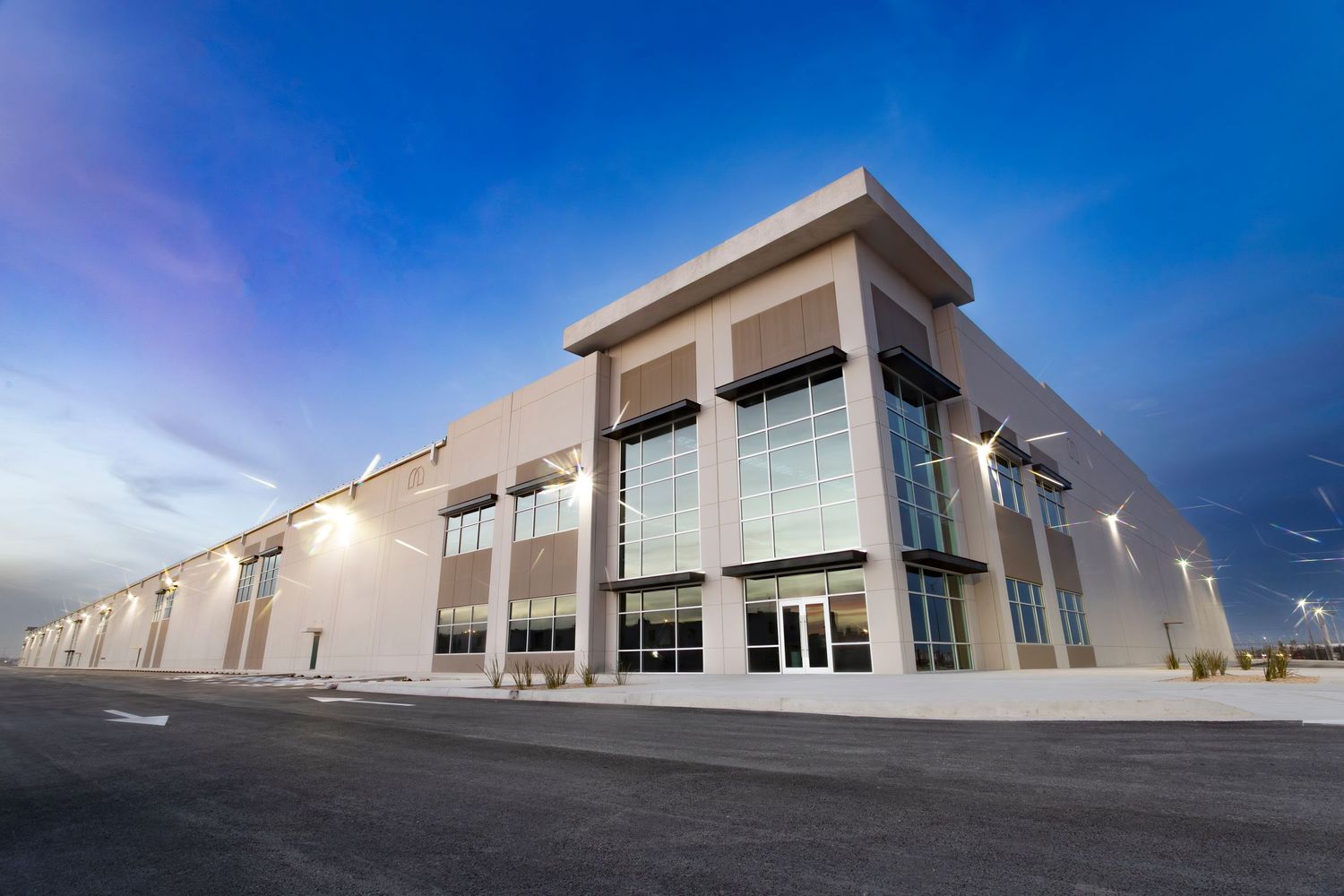 Modern industrial warehouse buildings with glass windows and loading docks under bright blue sky.