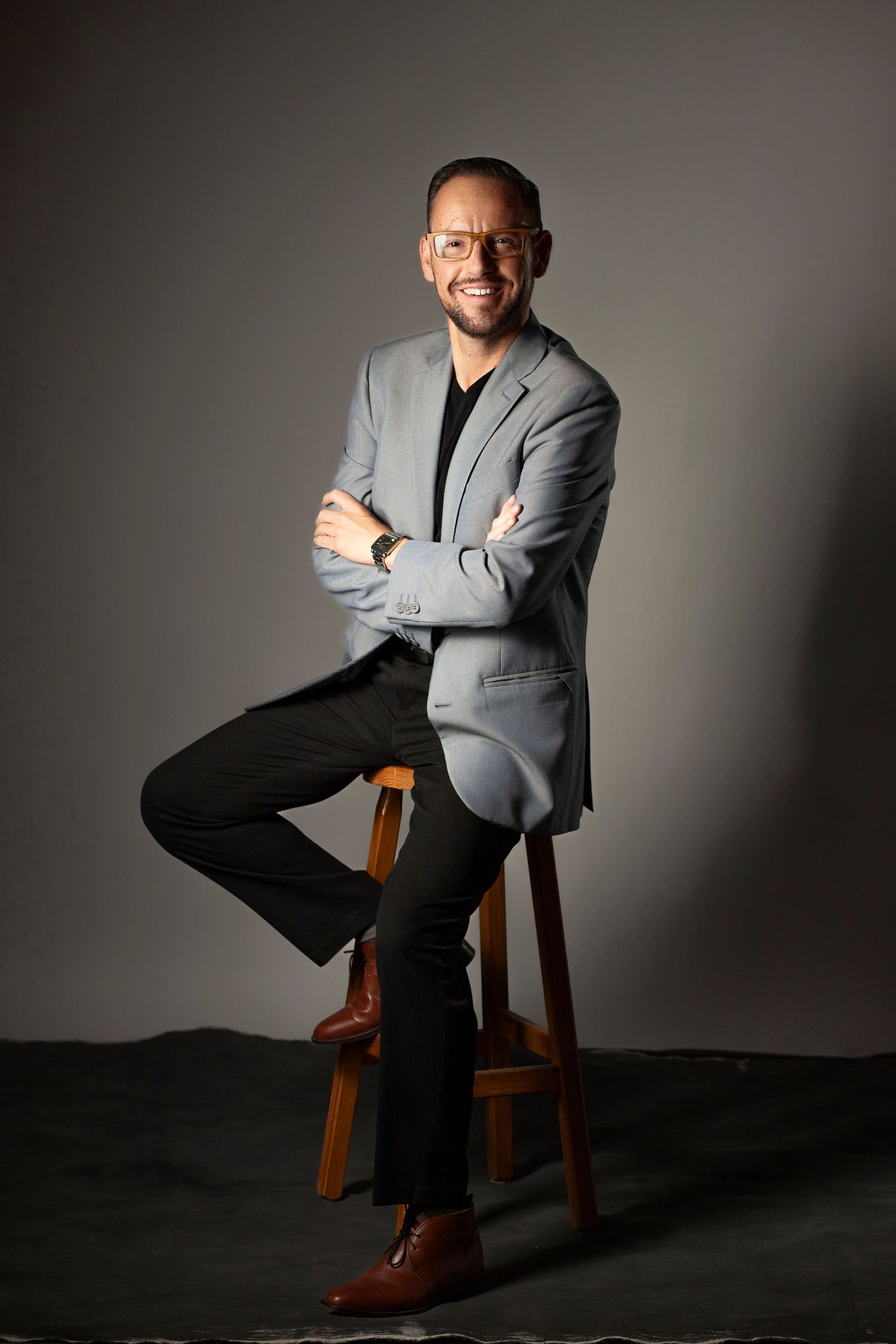 Professional studio portrait of someone seated on a wooden stool wearing a gray blazer, black shirt and brown shoes.