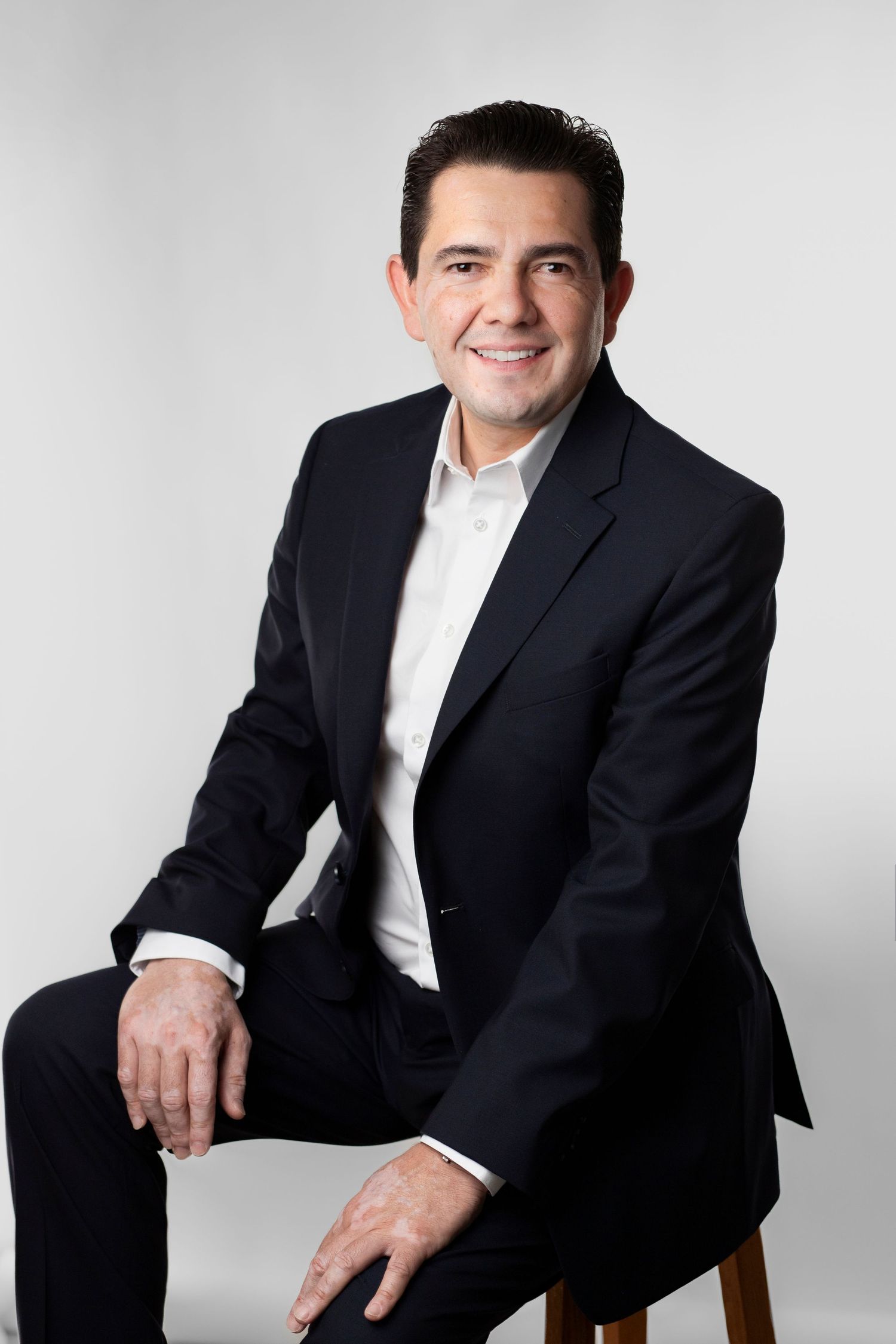 Professional studio portrait of person in black suit and white shirt seated in dynamic poses against light background.