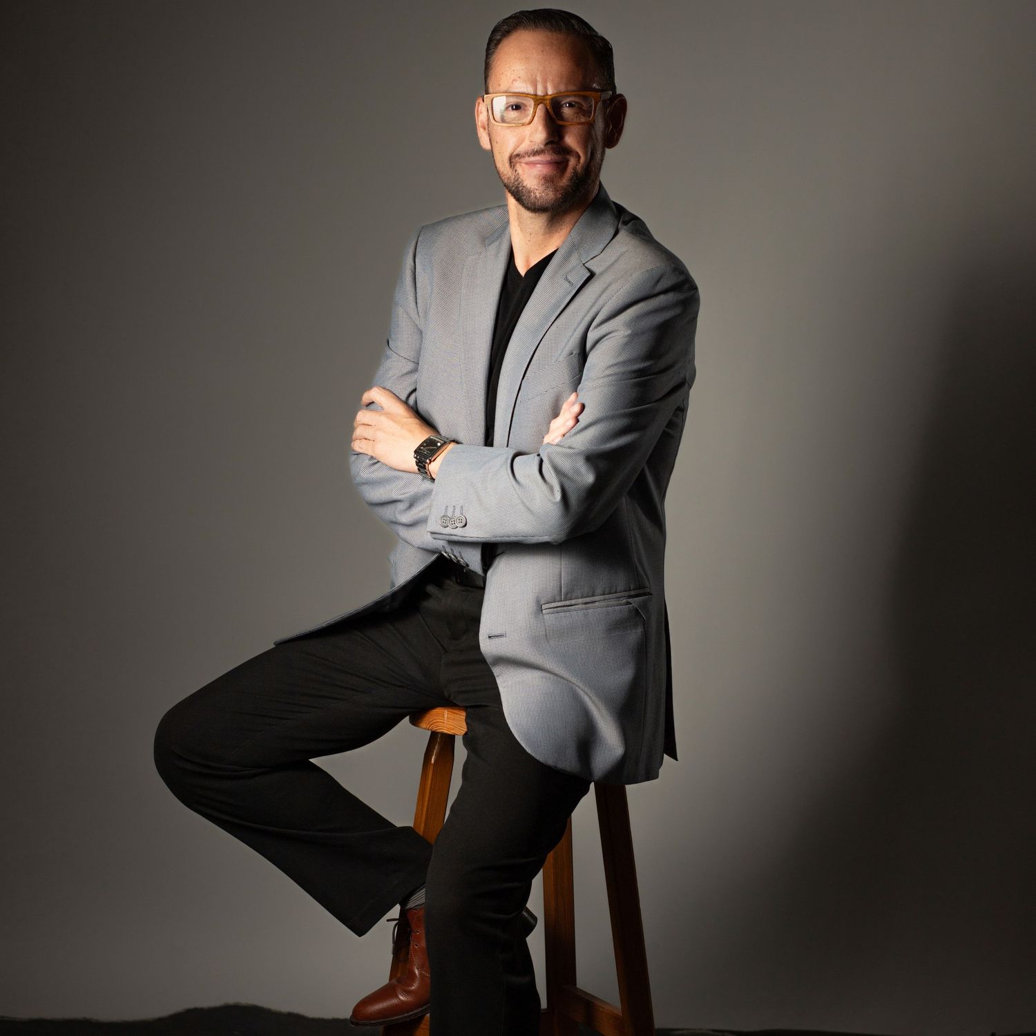 Professional studio portrait of someone seated on a wooden chair in a gray suit against a dark background.