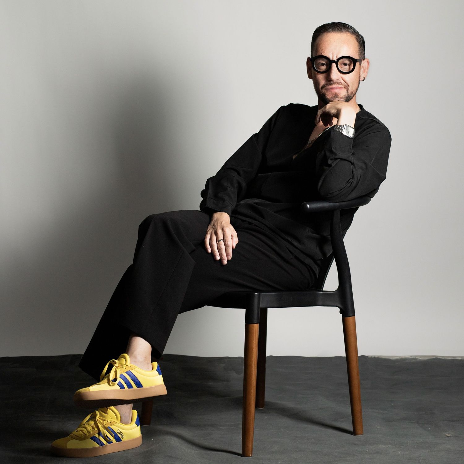 Professional studio portrait of person in black outfit and glasses sitting on wooden chair wearing yellow sneakers.