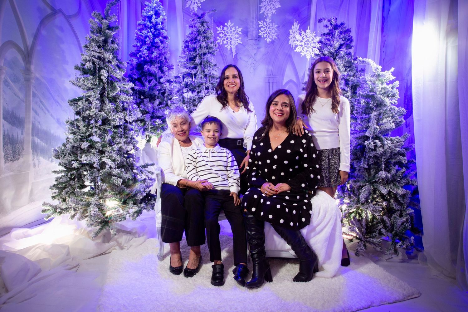 A group poses for a holiday portrait in a winter wonderland setting with frosted Christmas trees and purple lighting.