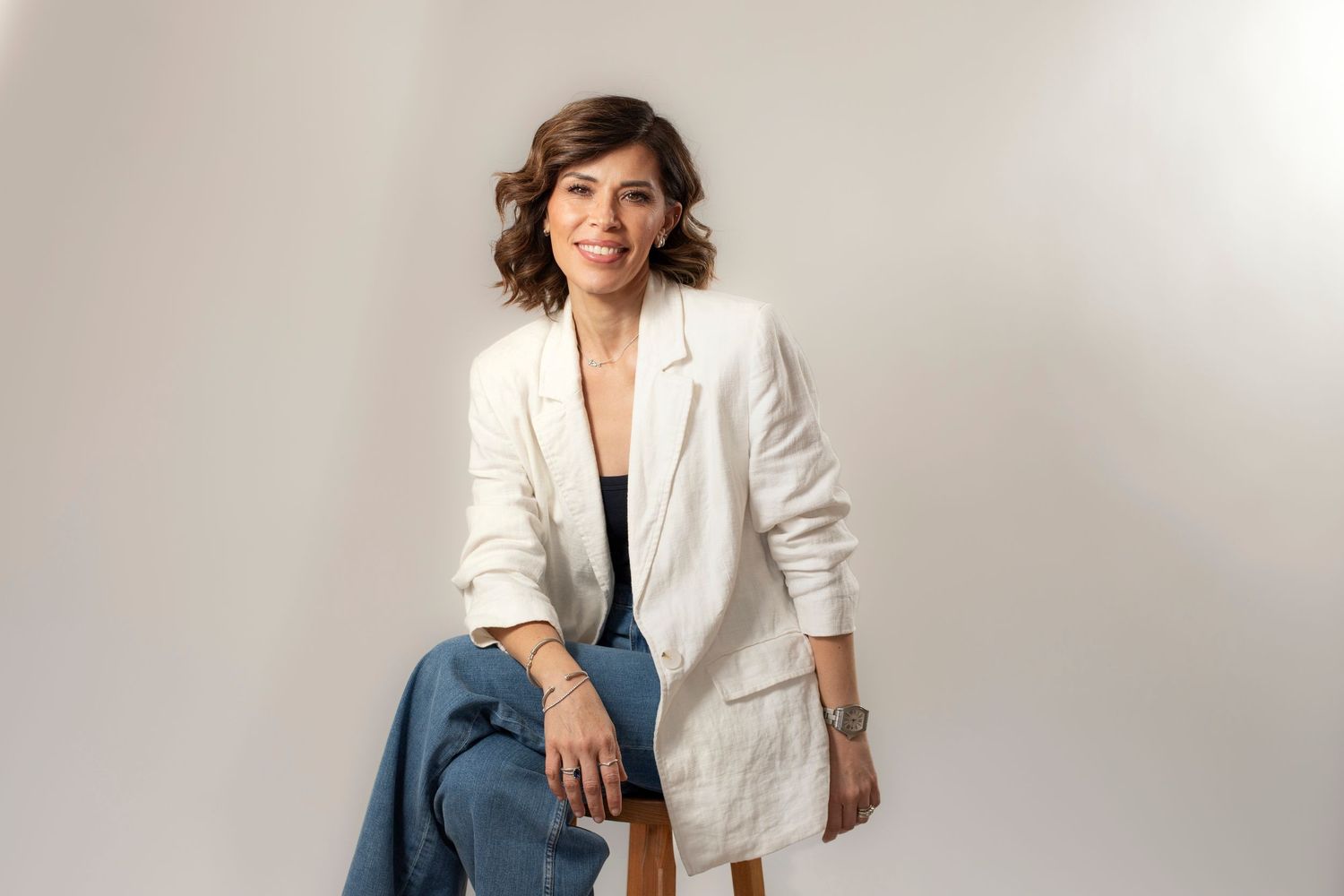 Woman in white blazer and jeans sits confidently on a stool against a light gray background.