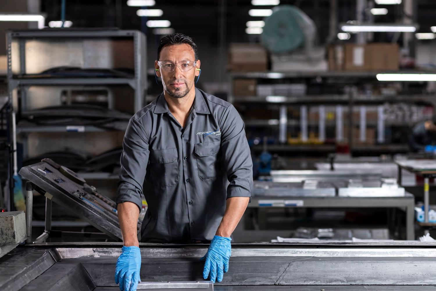 Factory worker in gray shirt and blue gloves stands confidently at industrial workbench in manufacturing facility.