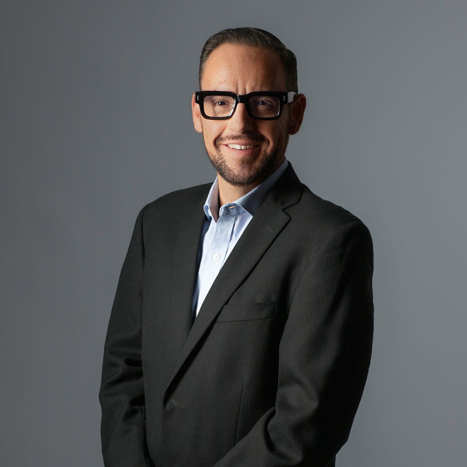 Professional wearing suit jacket, light blue shirt and glasses against grey studio backdrop.