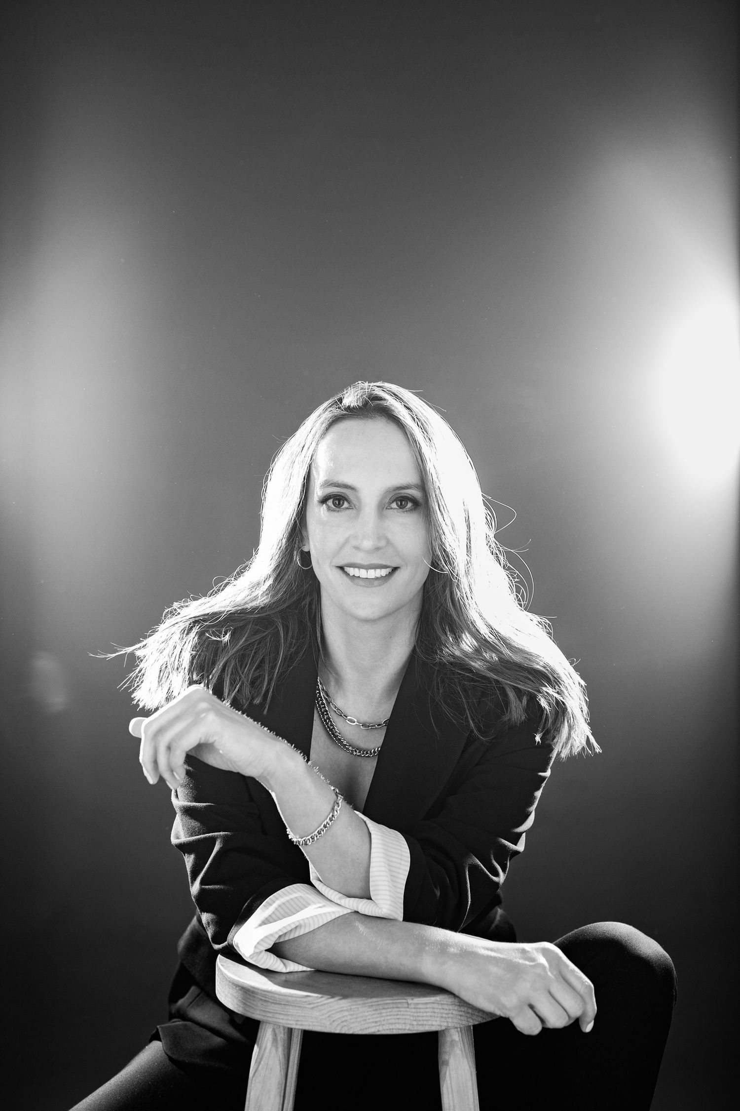 Black and white portrait of a smiling woman with long hair, leaning on a stool in a blazer with studio lighting.