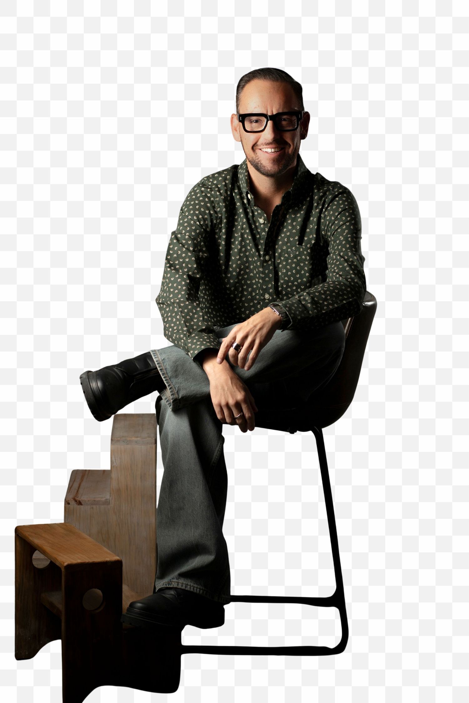 A person wearing glasses and a polka dot shirt sits on a wooden chair with a black metal frame.