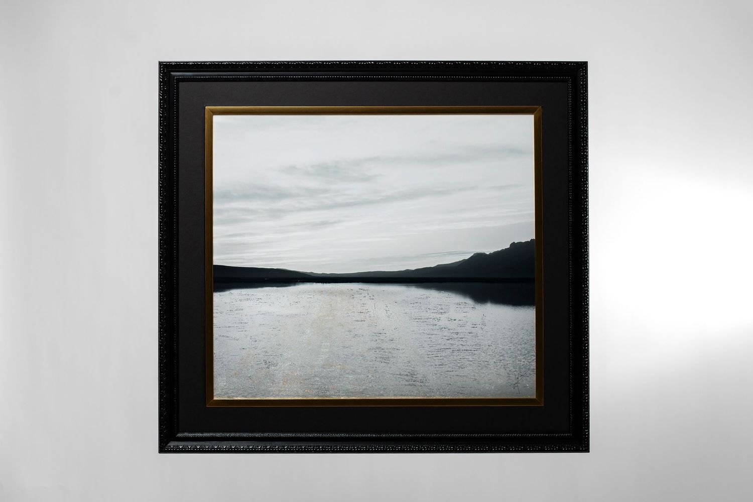 Framed landscape photograph of a calm lake reflecting a cloudy sky, displayed in a black and gold frame on a white wall.