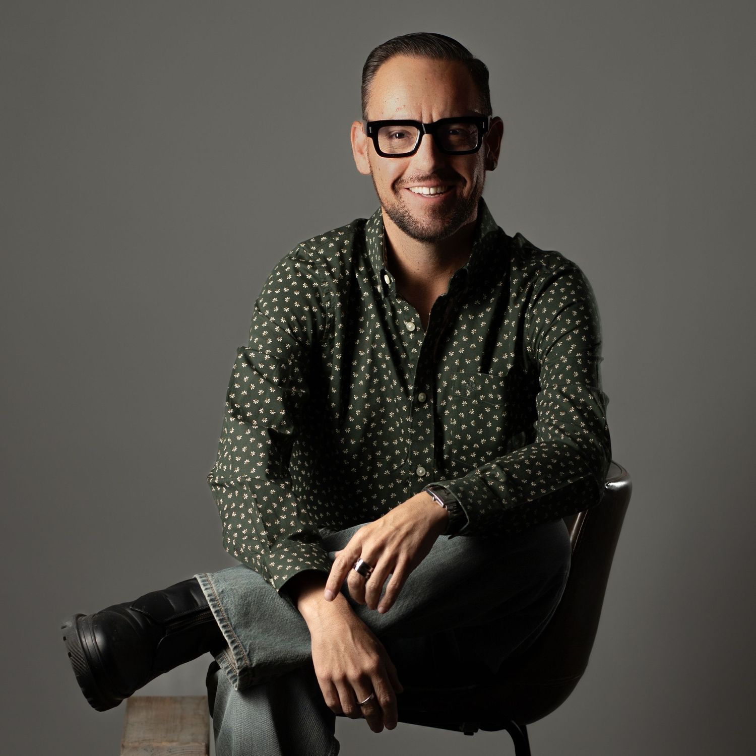 Professional portrait of someone in a polka dot shirt and glasses sitting casually in a modern chair against gray background.