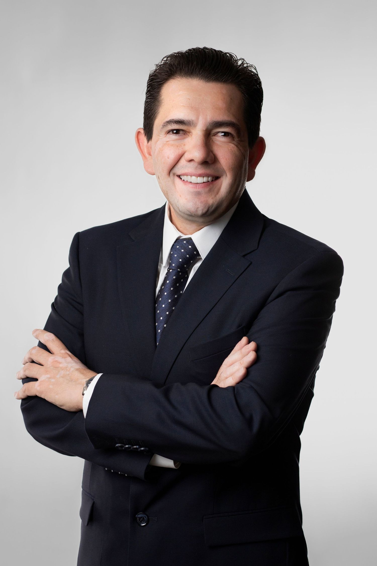 Professional wearing dark suit and polka dot tie poses with crossed arms against light studio background.