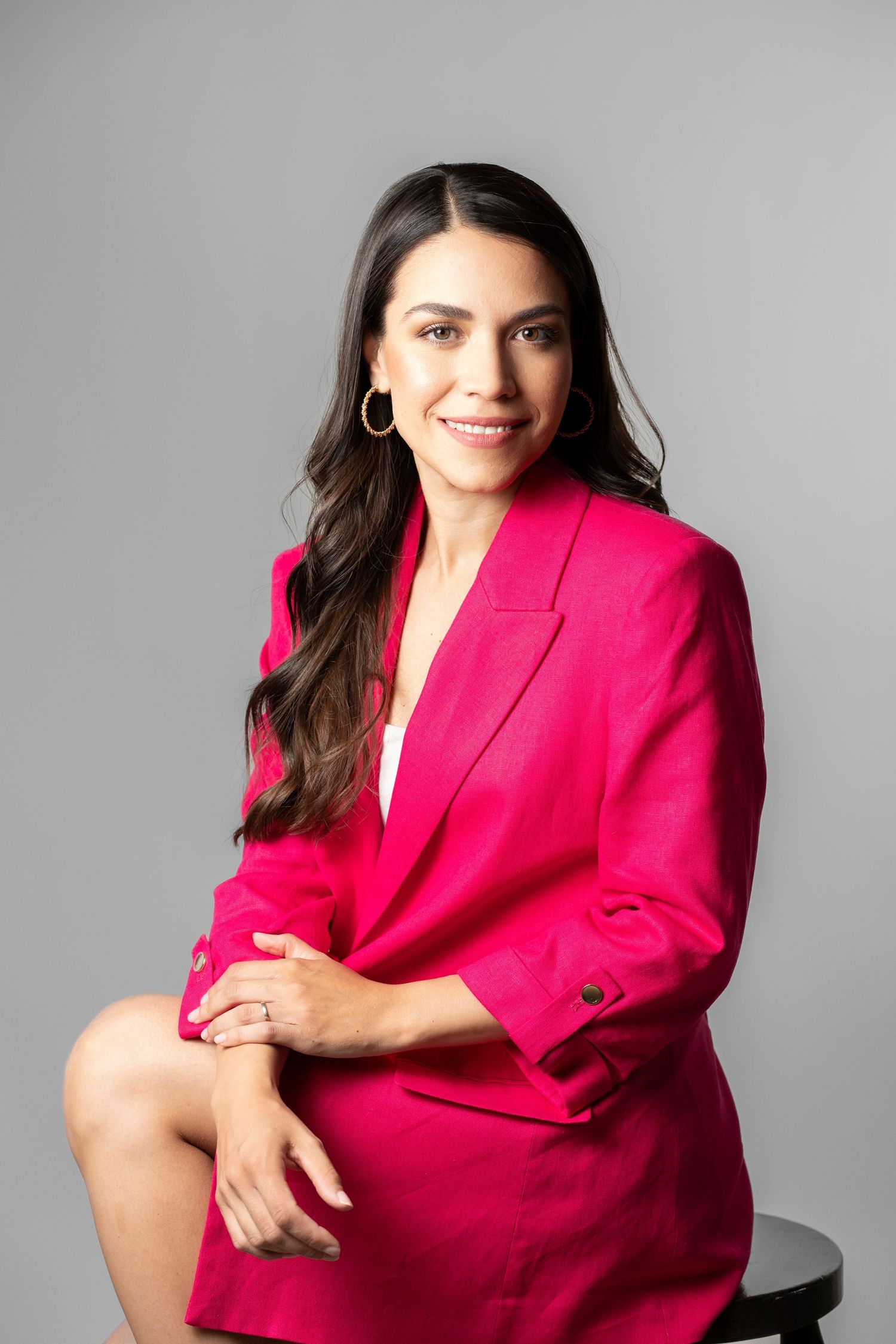 Professional studio portrait of someone wearing a vibrant hot pink blazer while seated against a gray background.