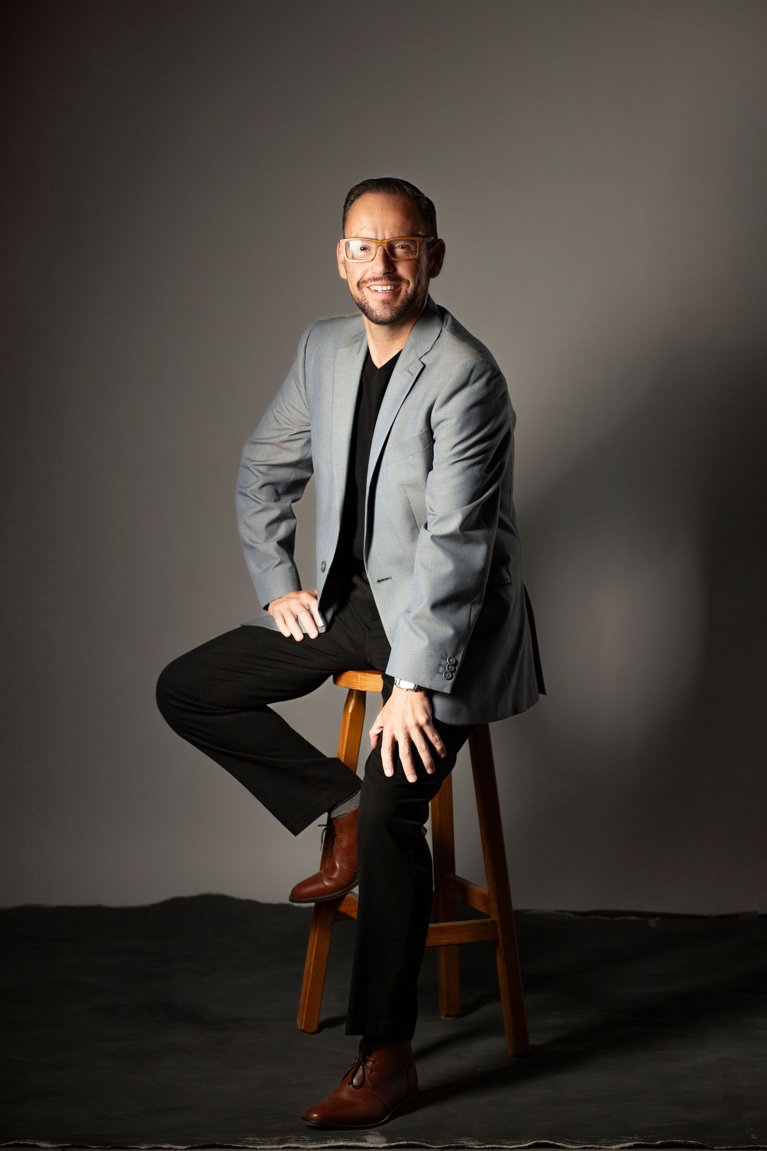 Professional studio portrait series showing seated figure in gray blazer and black pants against dark background.
