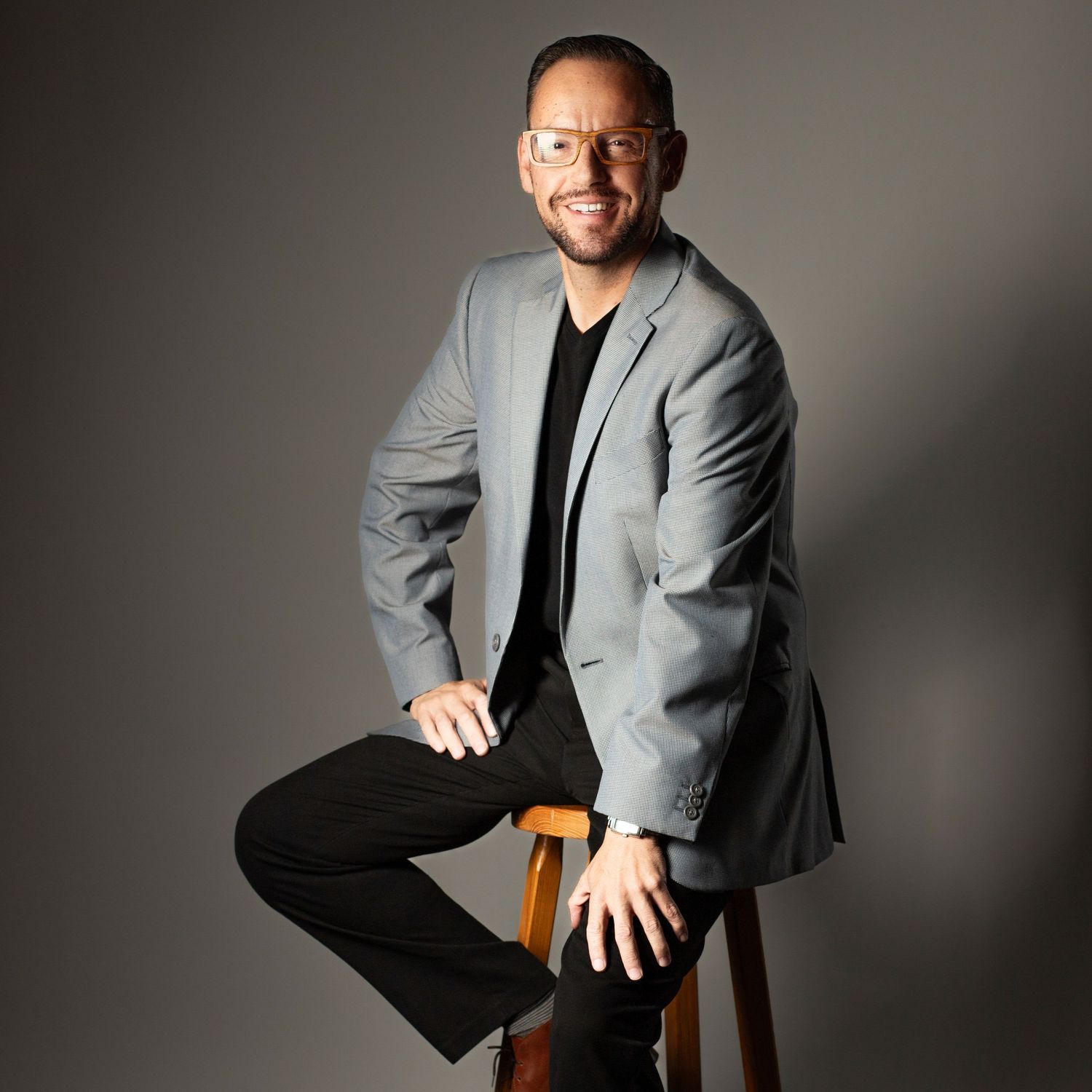 Professional studio portraits of someone in a gray blazer and black shirt sitting on a wooden stool against a gray backdrop.