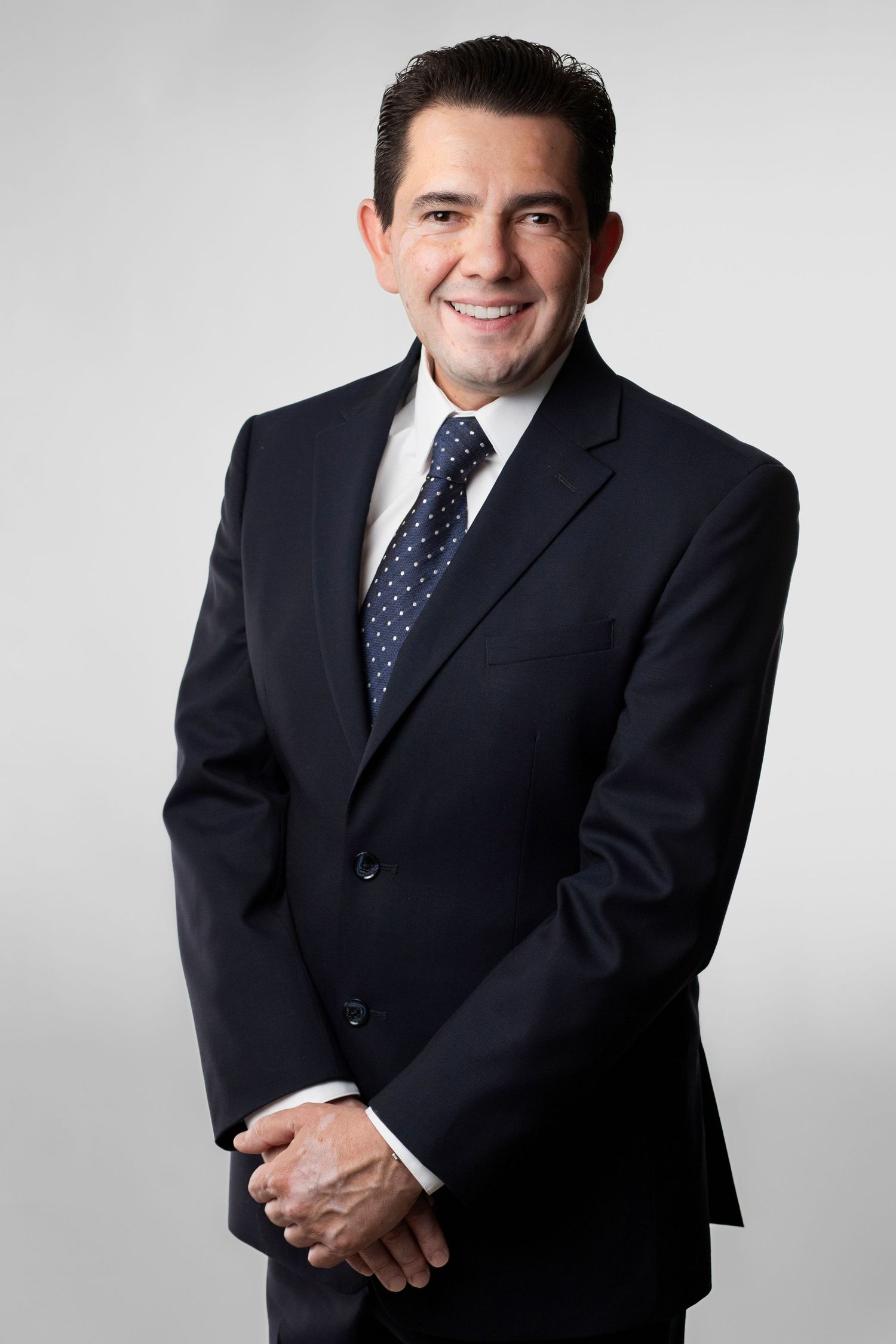 Professional headshot series of a person in a dark suit and polka dot tie against a light gray background.