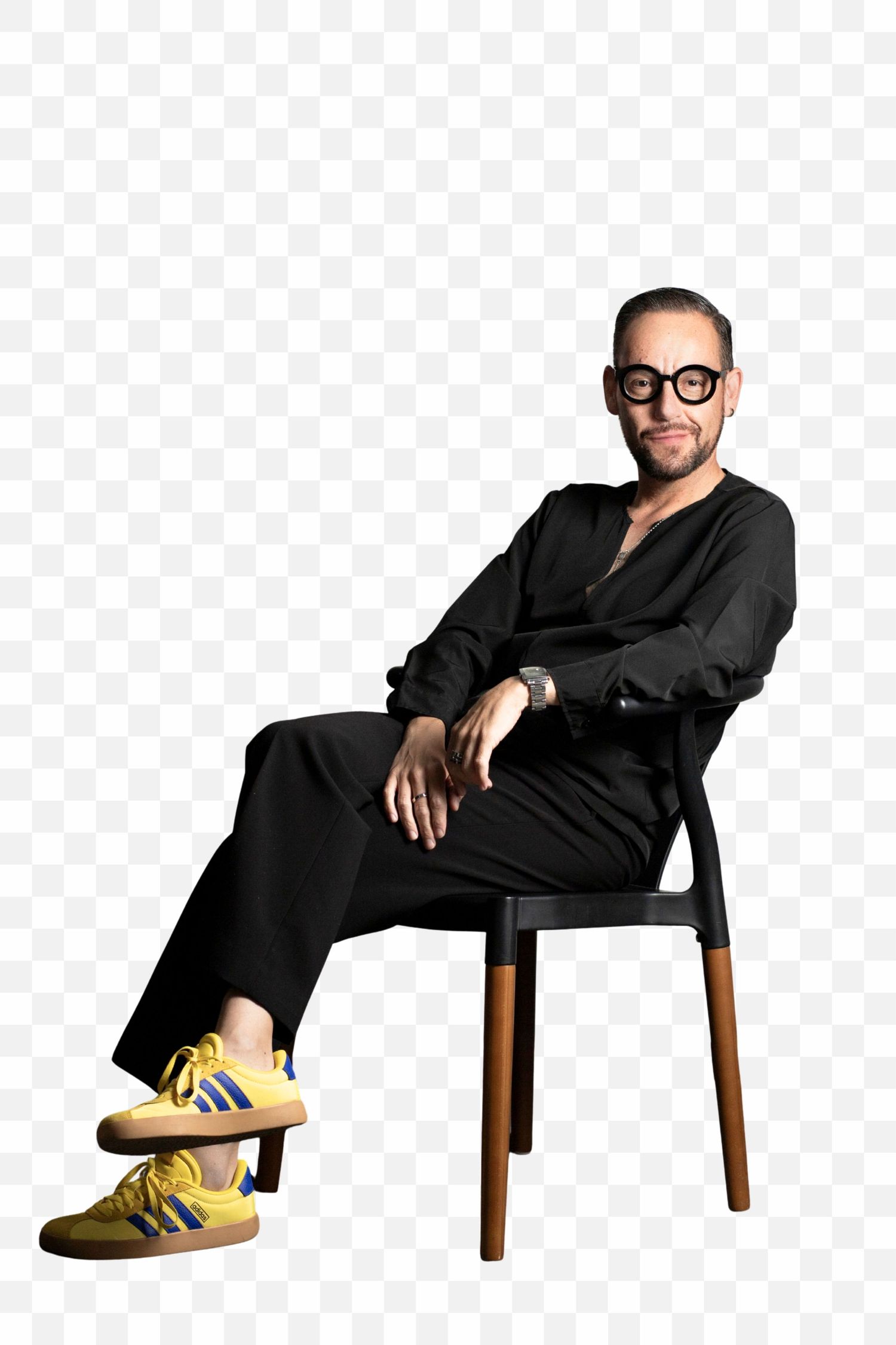 A person in black attire sits casually on a wooden chair wearing distinctive yellow sneakers and round glasses.