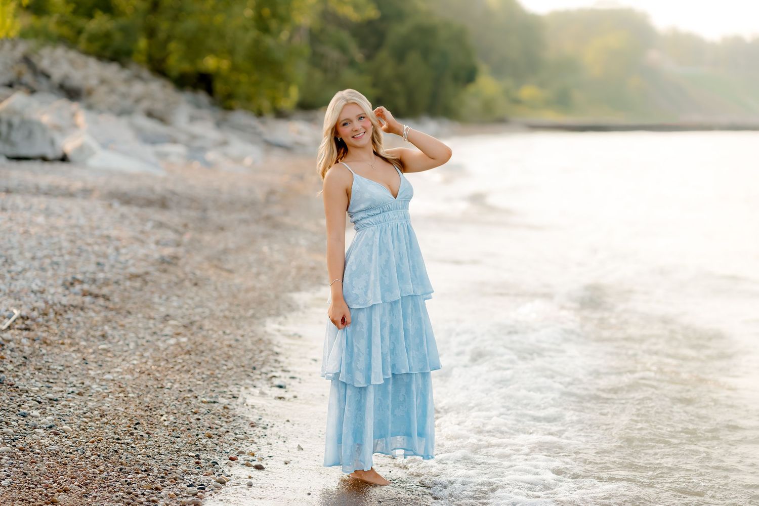 A blonde woman in a light blue tiered maxi dress stands barefoot on a rocky shoreline with trees in the background.