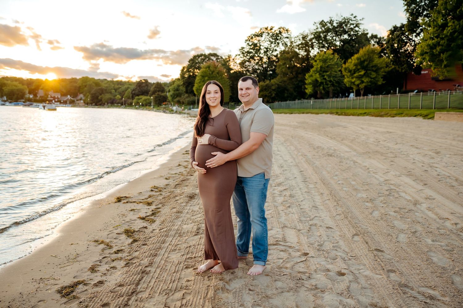 A couple embraces on a sandy beach at sunset with trees and water in the background.