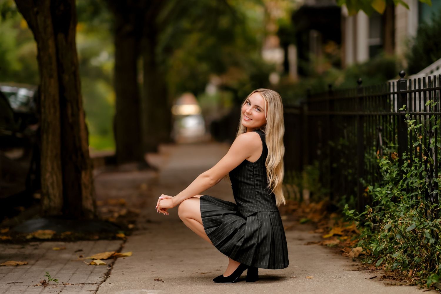 A person in a black dress crouches on a tree-lined sidewalk during autumn.