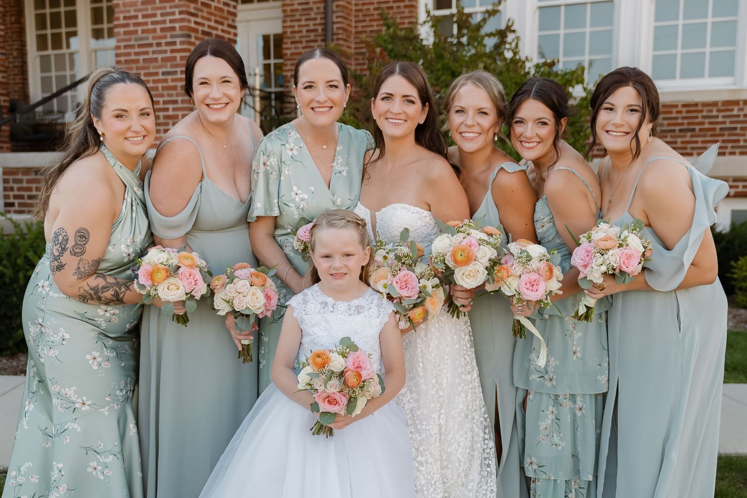 Bride in white gown poses with bridesmaids in sage green dresses and flower girl, holding pink and peach bouquets.