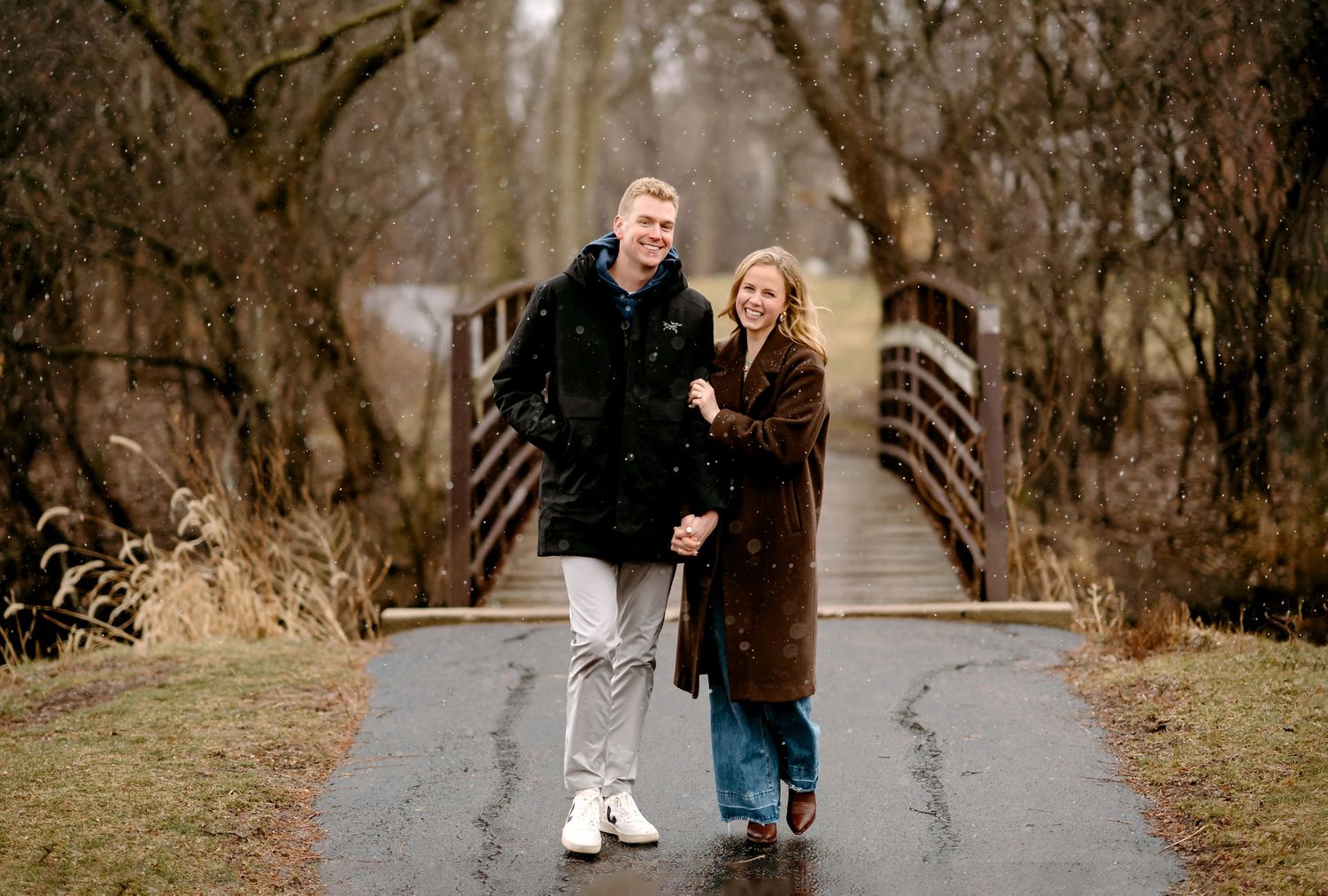 Couple smiling and holding hands on a rainy path near a wooden bridge surrounded by bare winter trees.