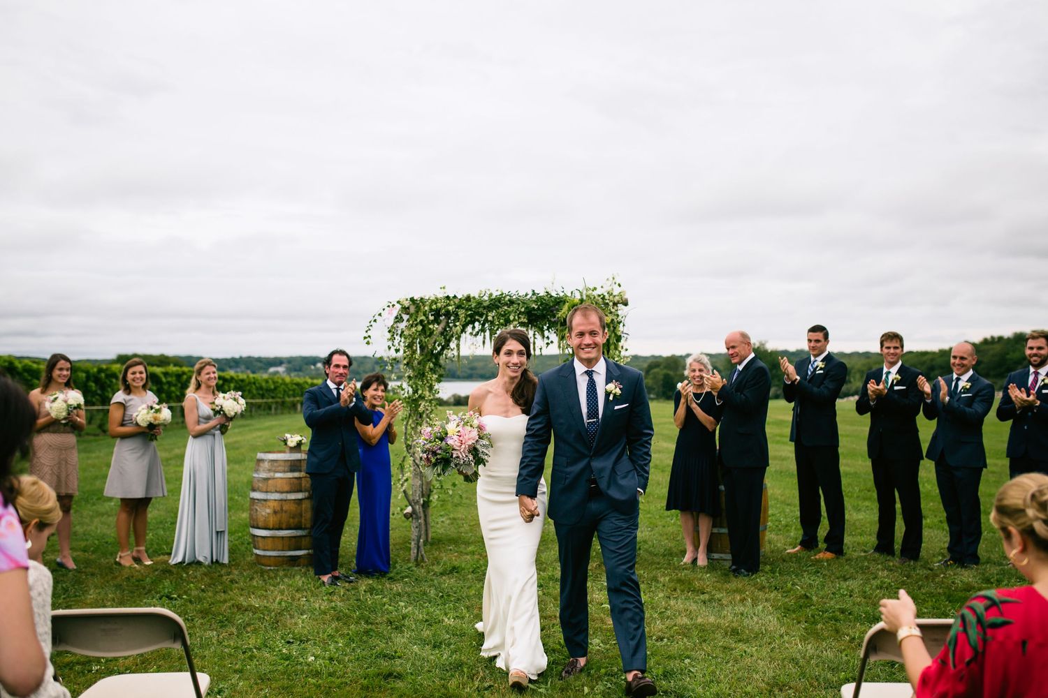 Wedding guests applaud during an outdoor ceremony as newlyweds celebrate on a grassy hilltop.
