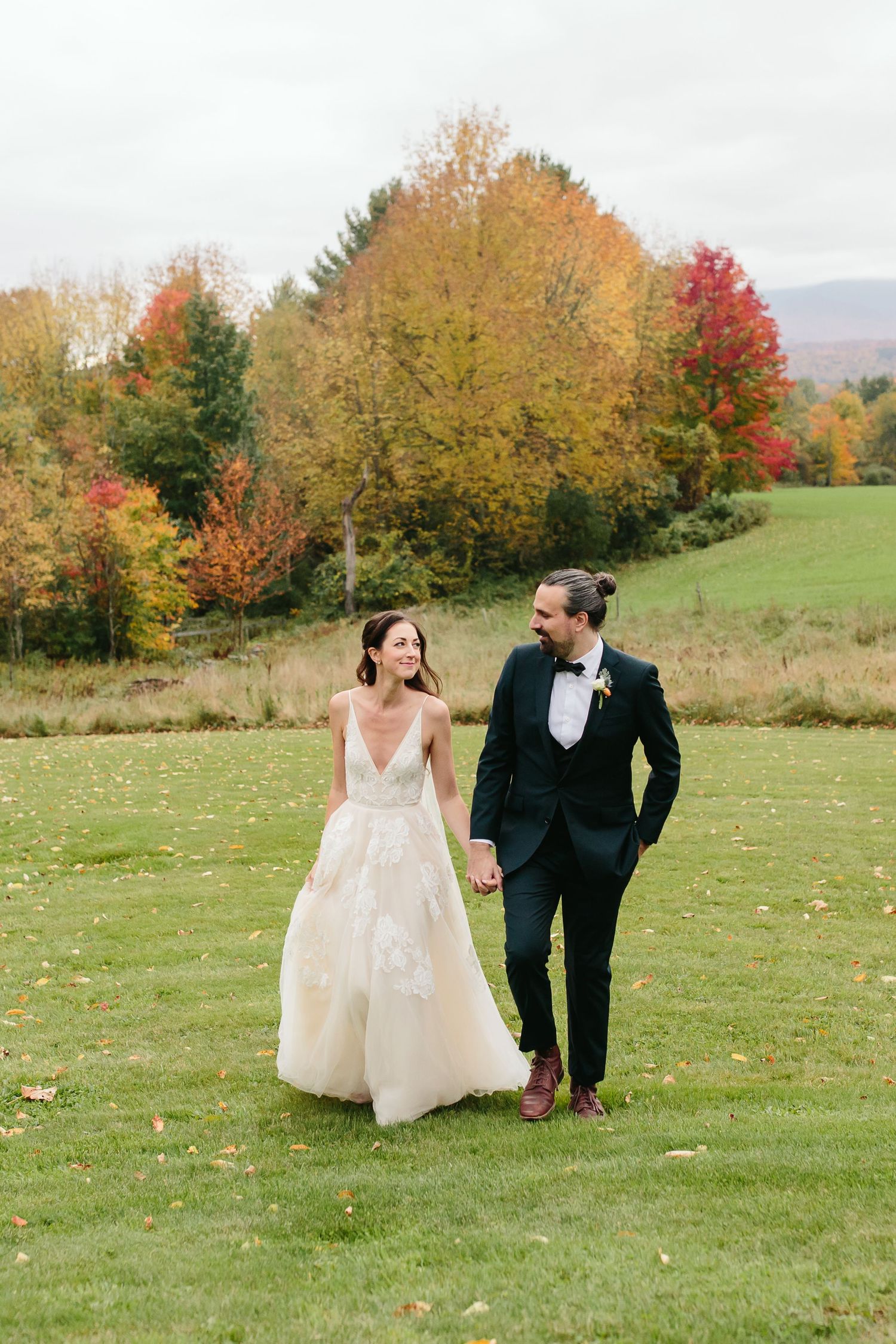 A couple walks hand in hand through a field with stunning fall foliage in the background.