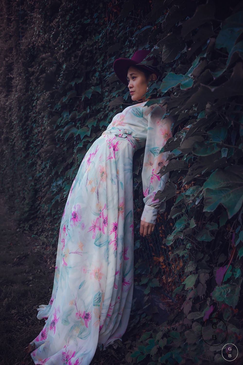 A figure in a flowing white floral dress poses dramatically against a dark garden wall with lush foliage.
