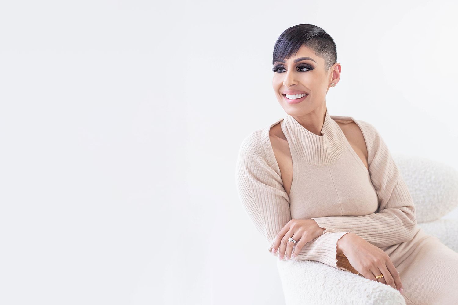 Someone in a beige sweater and halter top sits comfortably on a white couch while smiling at the camera.
