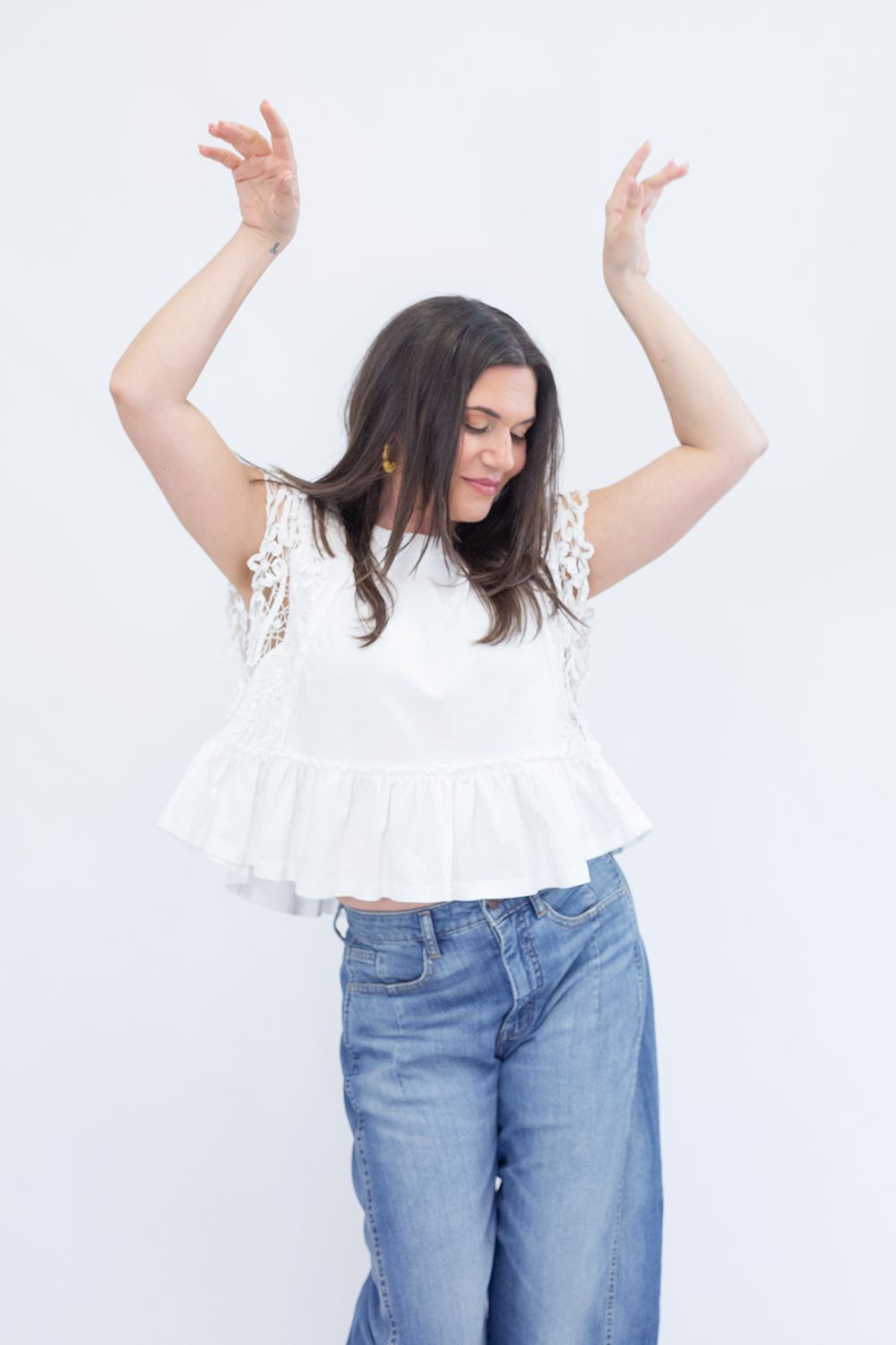 A person wearing a white eyelet top and blue jeans poses with raised arms against a white background.