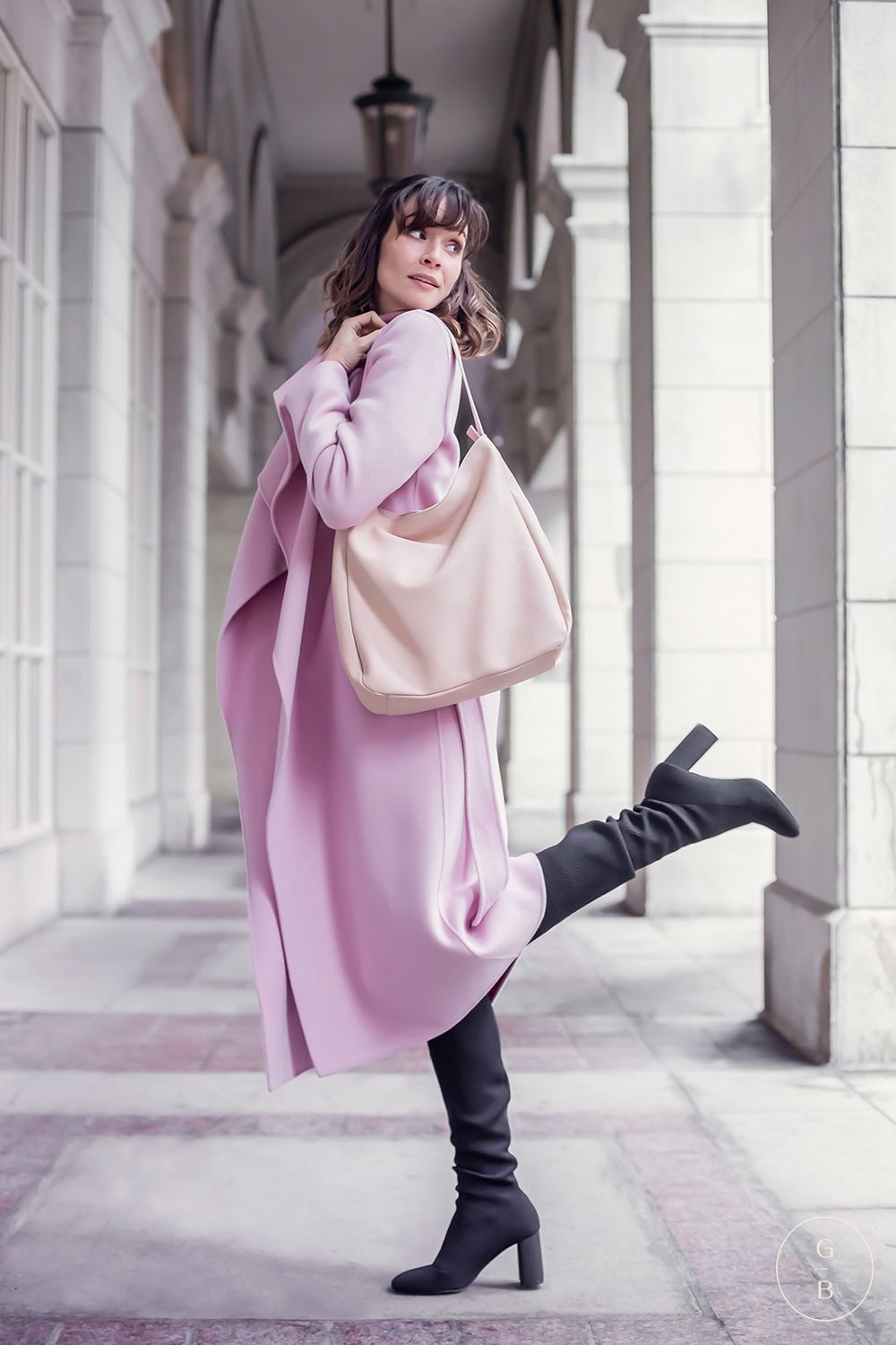 Fashion model in pink oversized sweater and black pants poses elegantly against white architectural backdrop.