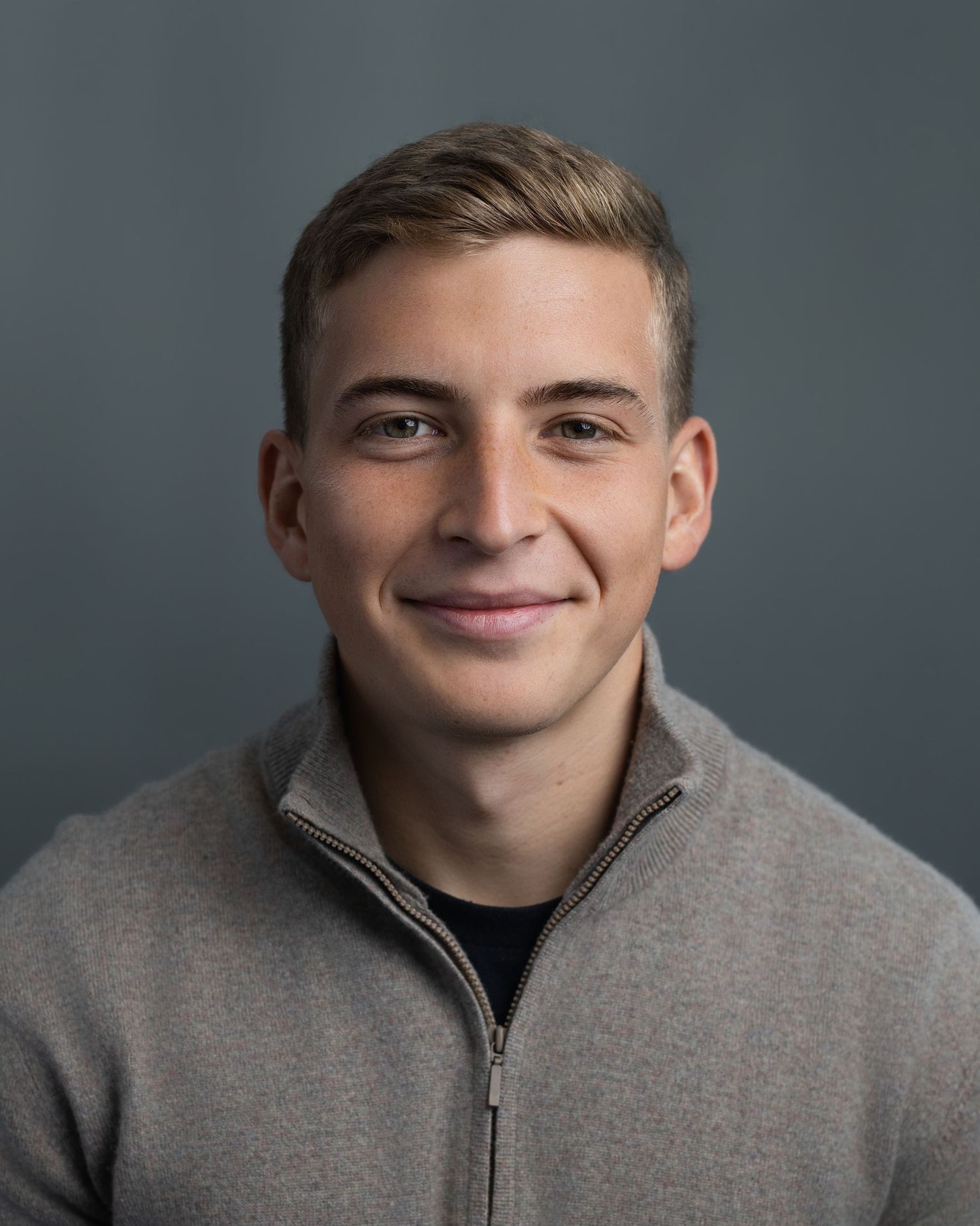 Young man with blonde hair smiling, wearing a gray zip-up sweater, professional headshot photo.