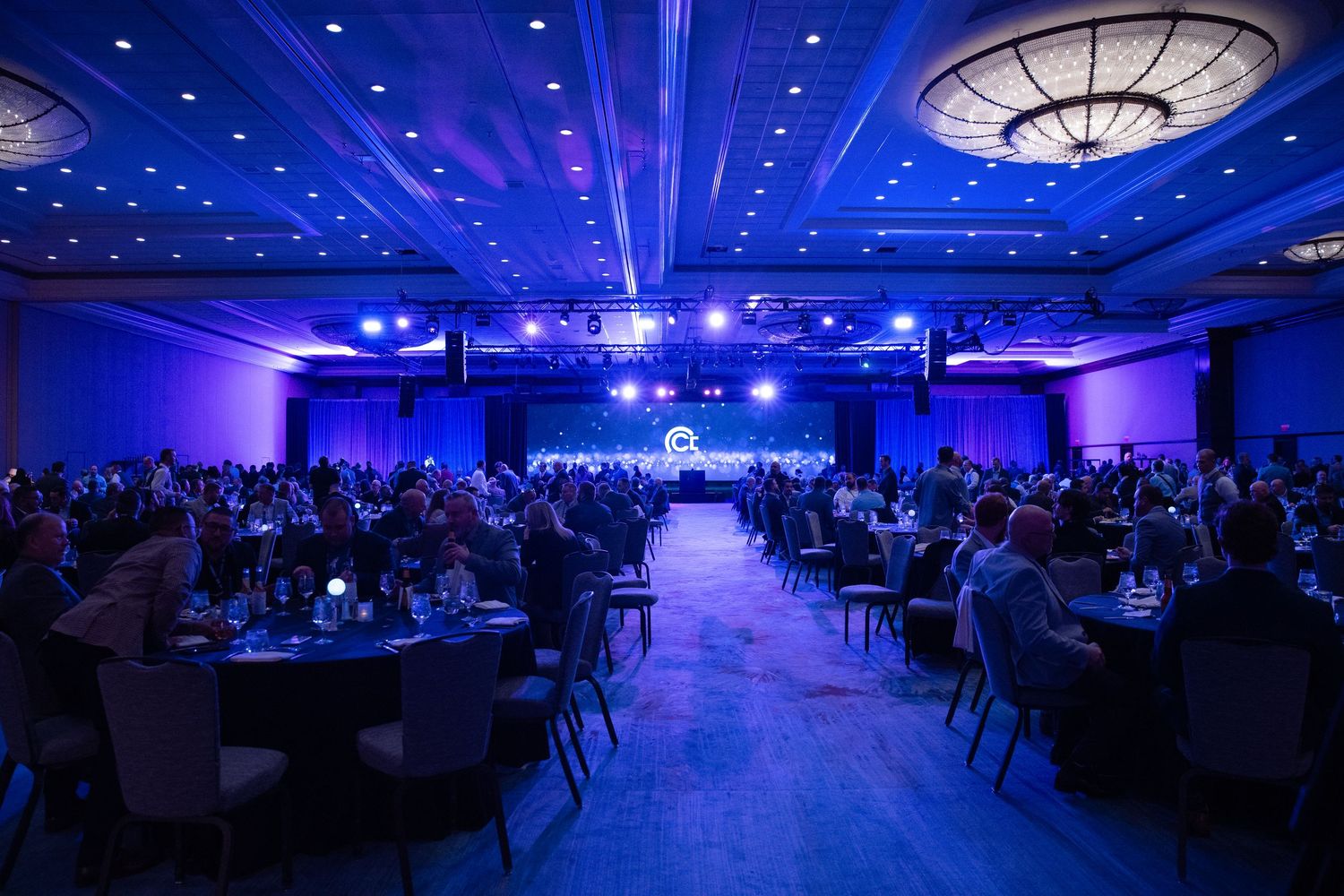 Large corporate gala event in a grand ballroom with blue lighting, round tables, and hundreds of attendees facing a stage.