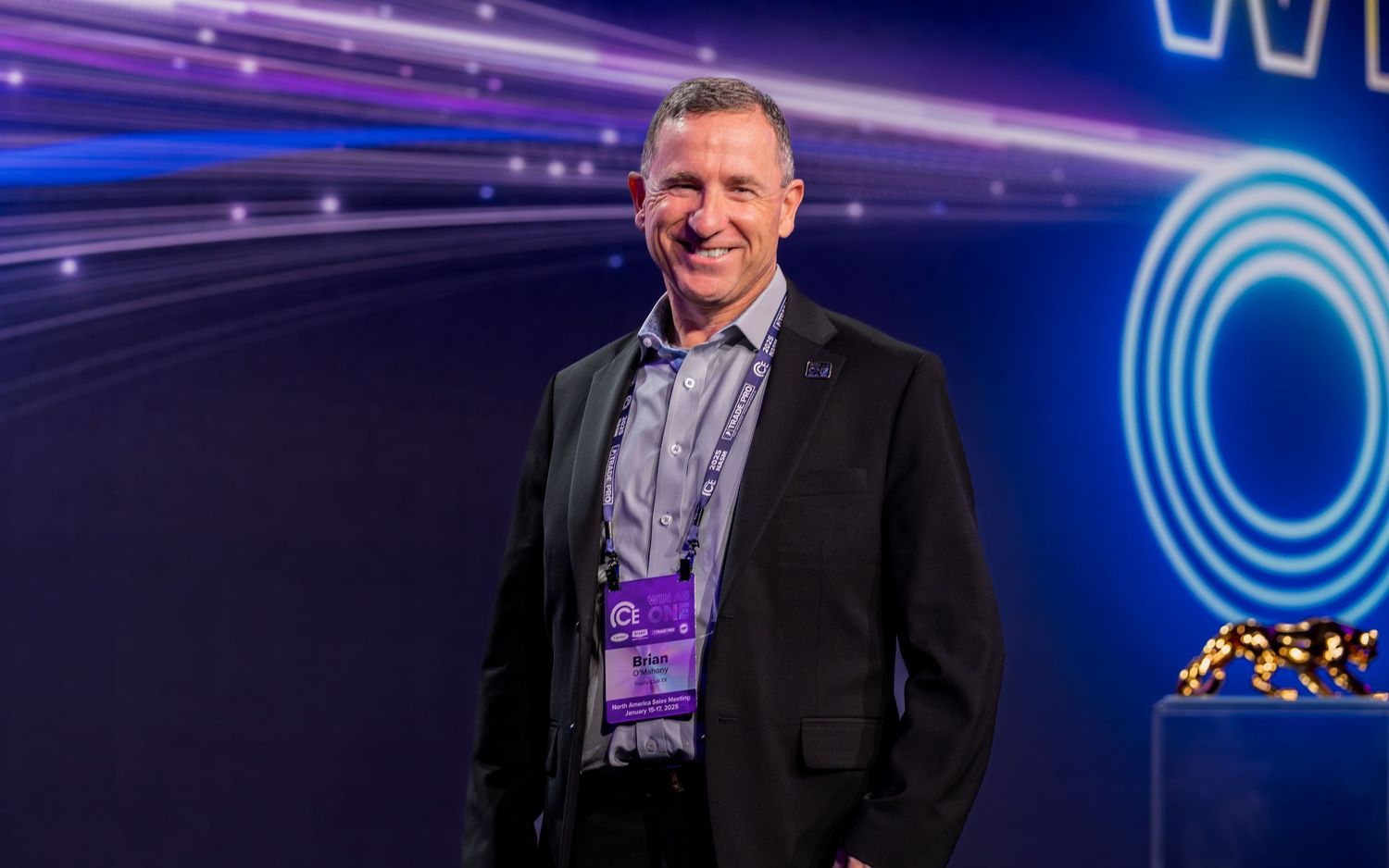 A smiling man in a black blazer and lanyard stands confidently in front of a vibrant purple tech event backdrop.