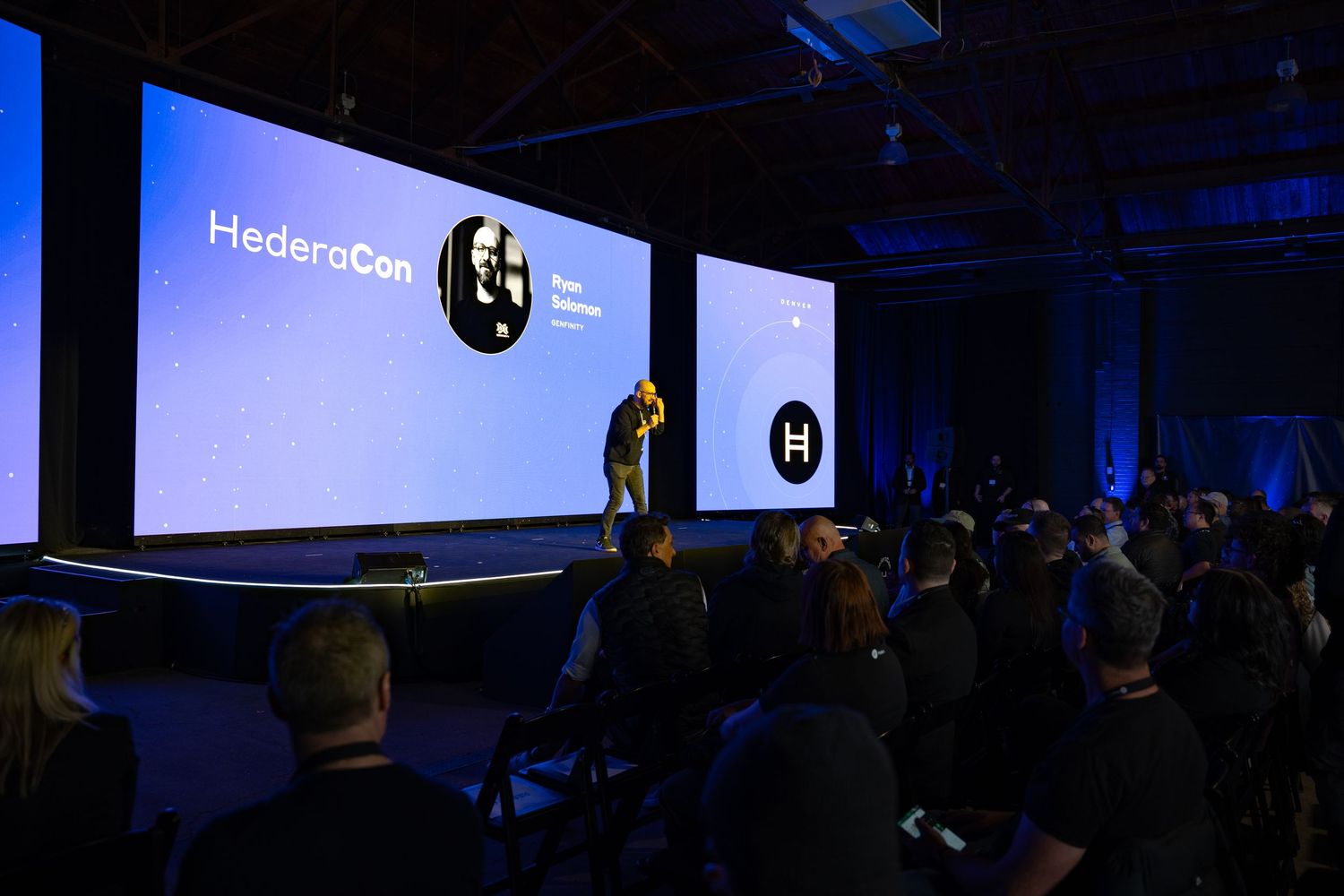 Speaker presents on stage at HederaCon conference with large screen displaying Hedera logo and branding.