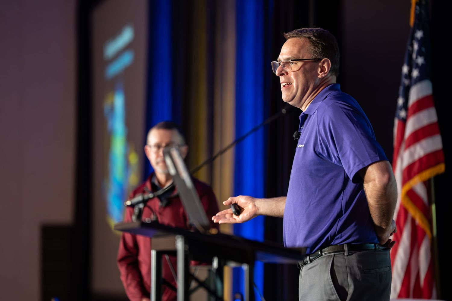 Man in blue polo shirt speaking at a podium on stage with an American flag nearby.