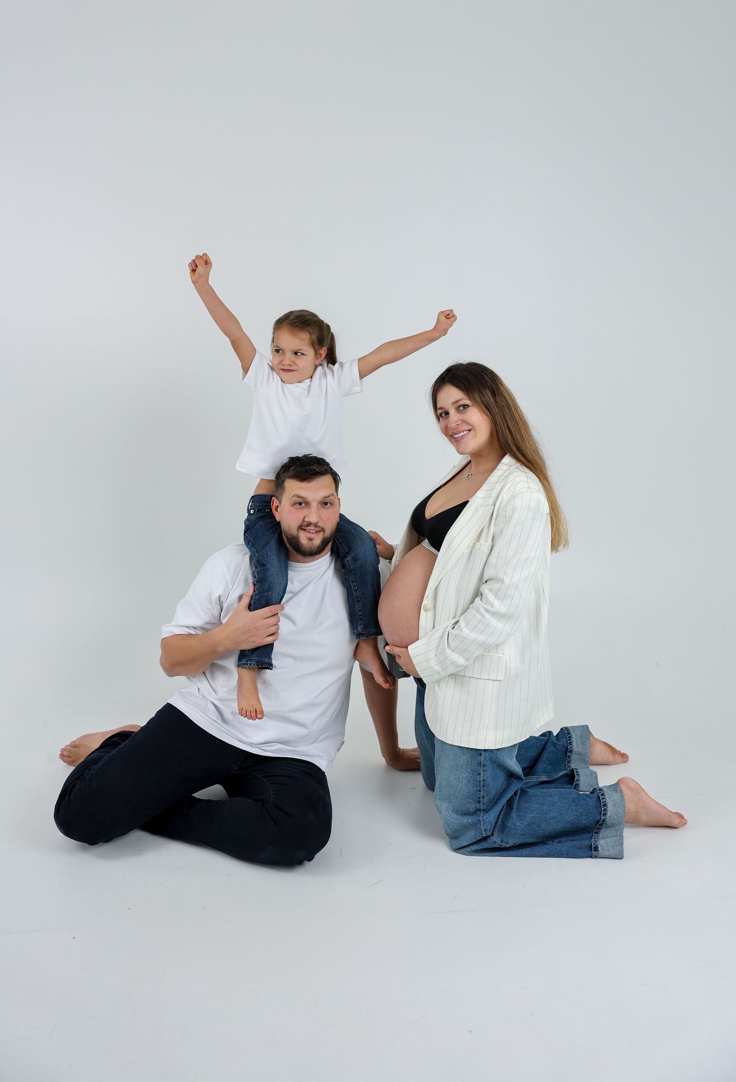 A happy family with a pregnant mother, father, and young child posing together on a white studio background.