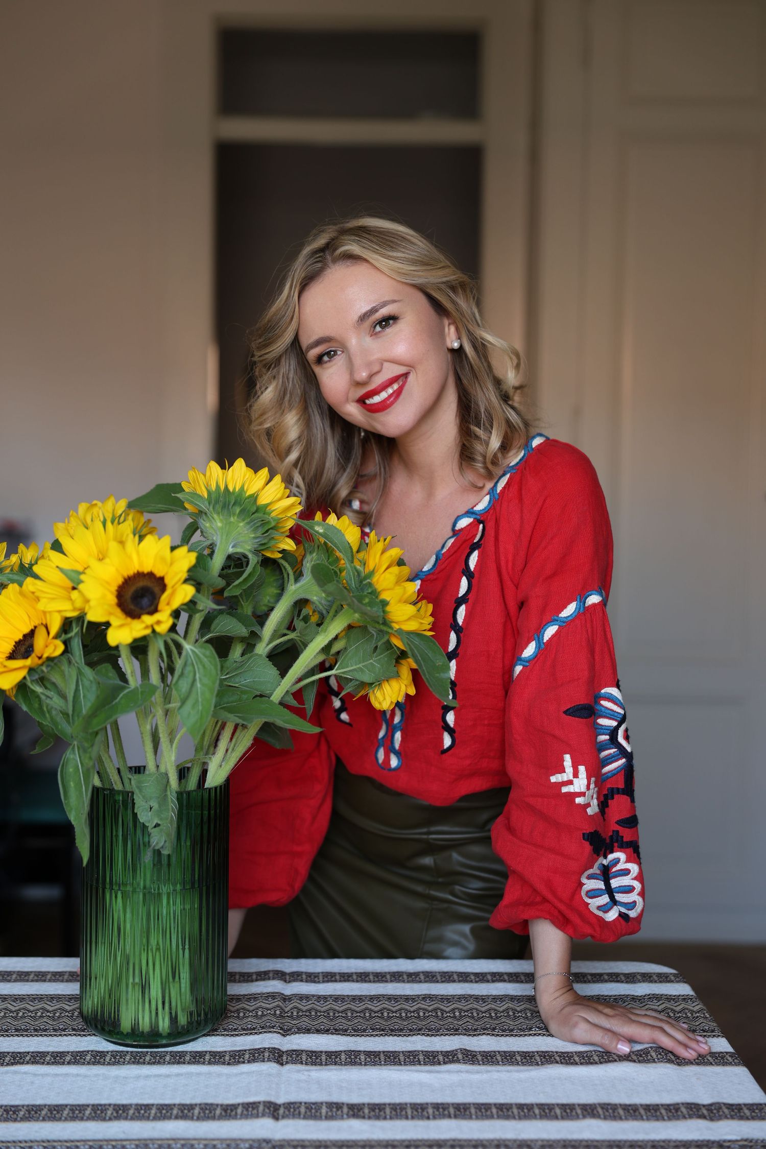 Smiling woman in red embroidered blouse with sunflowers in glass vase on striped tablecloth indoors.