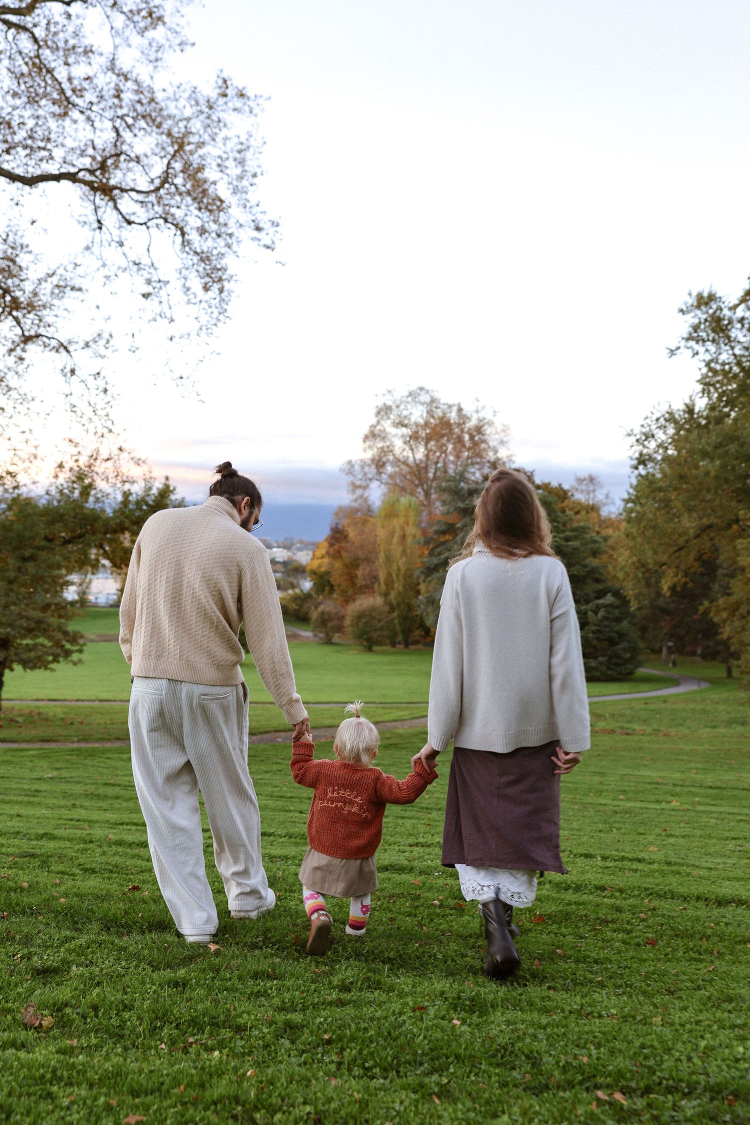 Family of three walking hand-in-hand across lush green park, two adults guiding toddler in red sweater on autumn day.