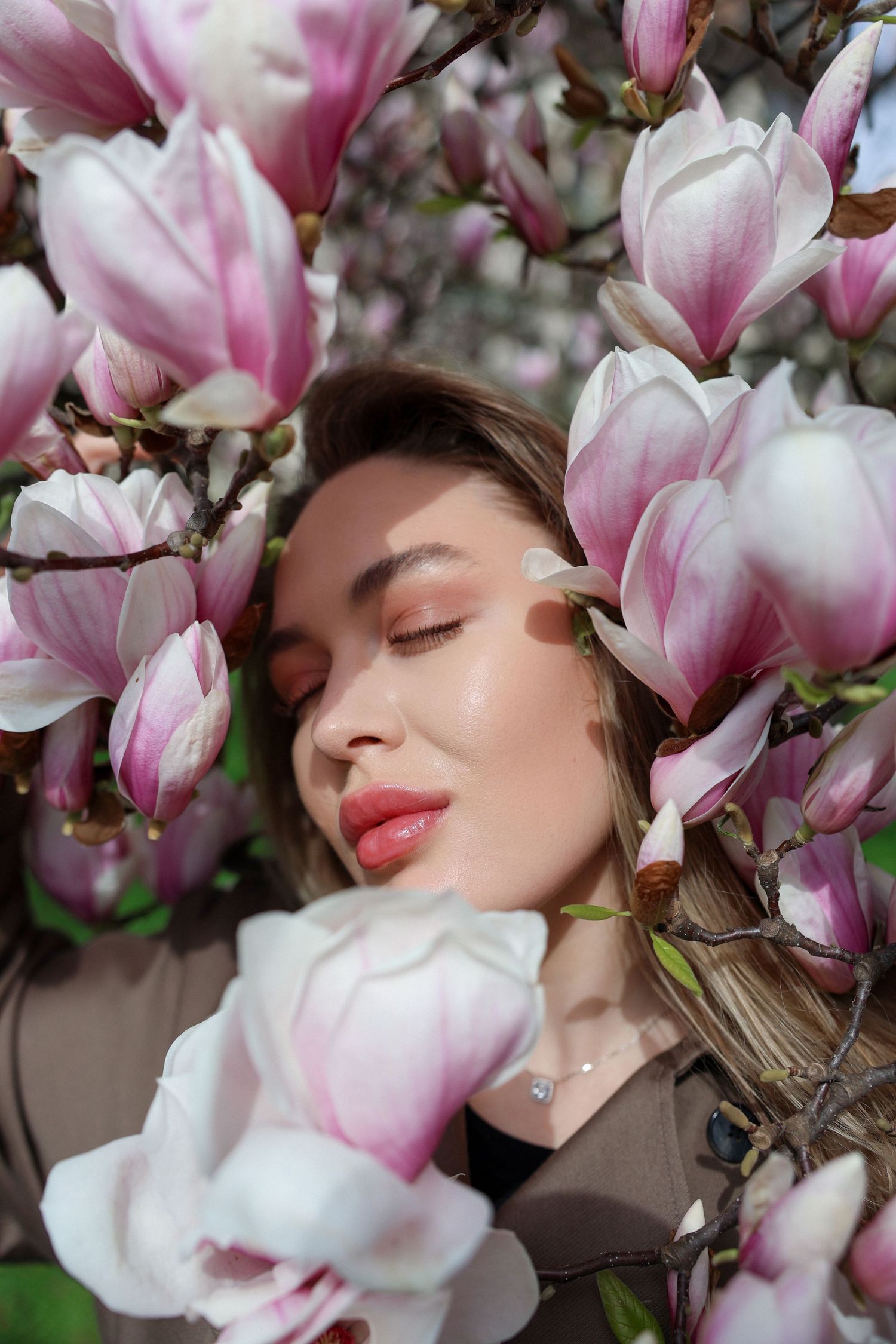 Young woman with glowing skin and pink lips surrounded by blooming pink magnolia flowers in soft spring light.