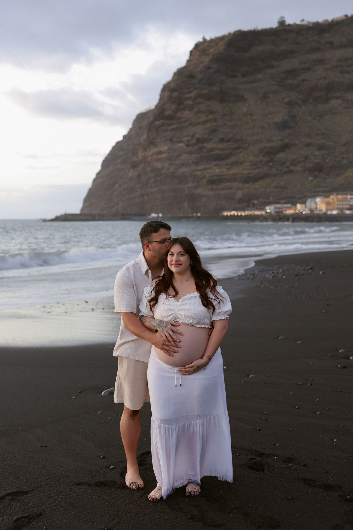 Couple on black sand beach, pregnant woman in white dress, man embracing her bump, dramatic cliff and ocean backdrop.