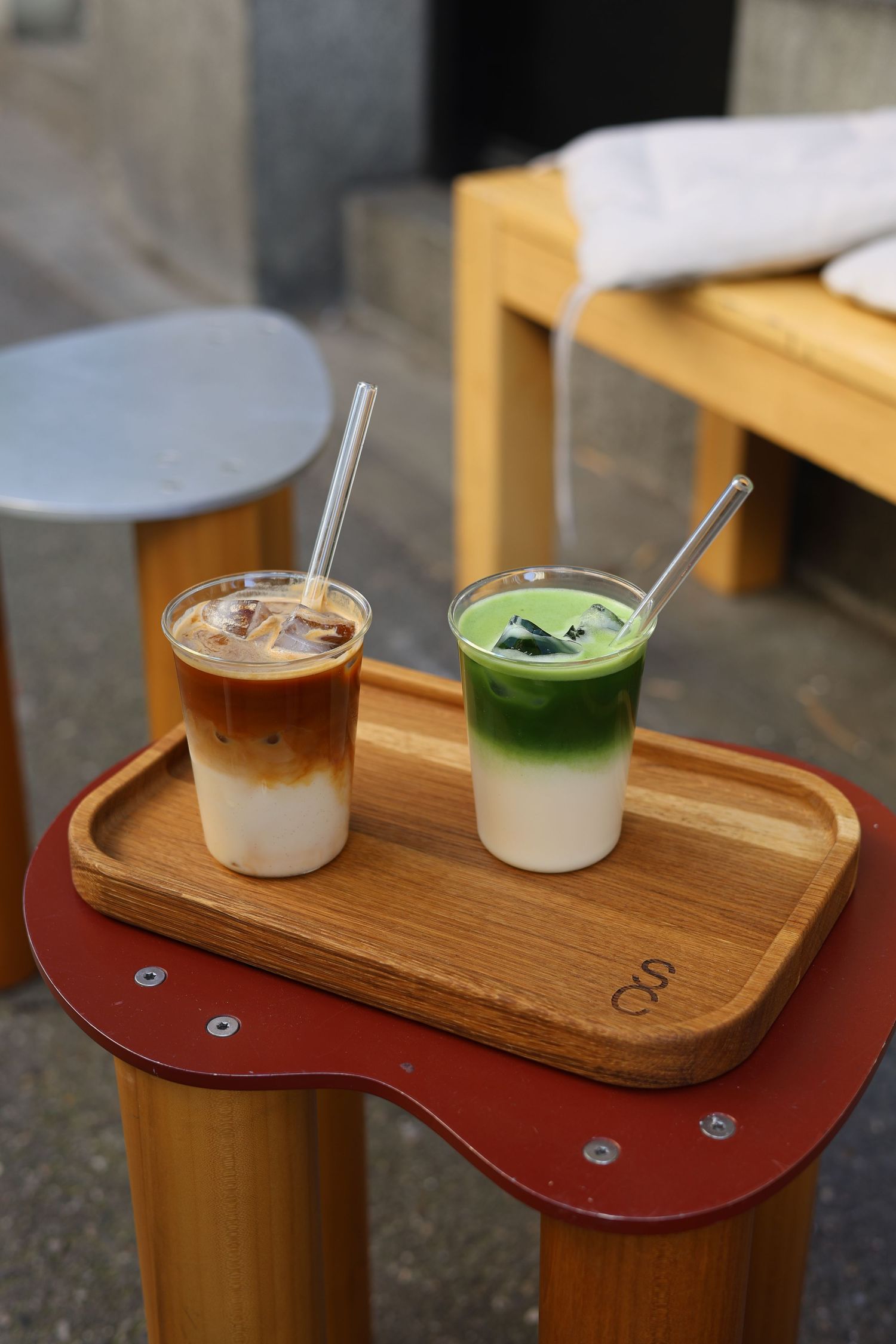 Two iced drinks, iced latte and matcha latte, served on a wooden tray at an outdoor cafe.