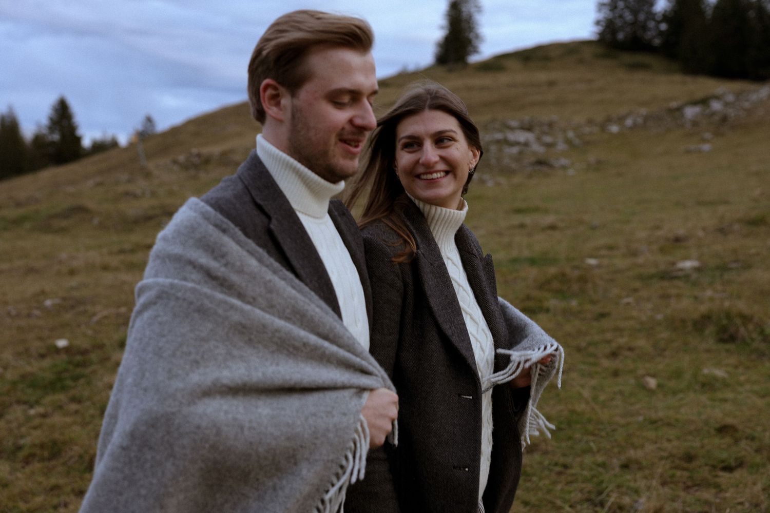 Couple wrapped in gray blanket smiling on a green hillside, wearing dark coats in an outdoor autumn setting.