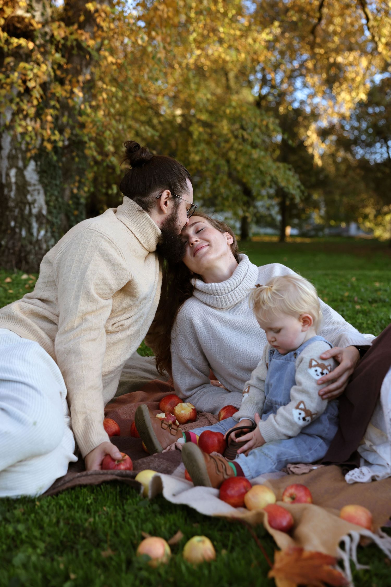 A young family enjoys an autumn picnic on a blanket with apples scattered around them in a park.
