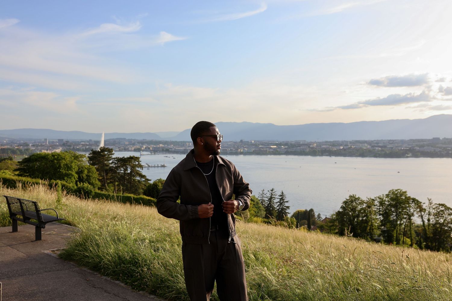 Man standing on a scenic overlook with a lake, city skyline, and lush greenery in the background.